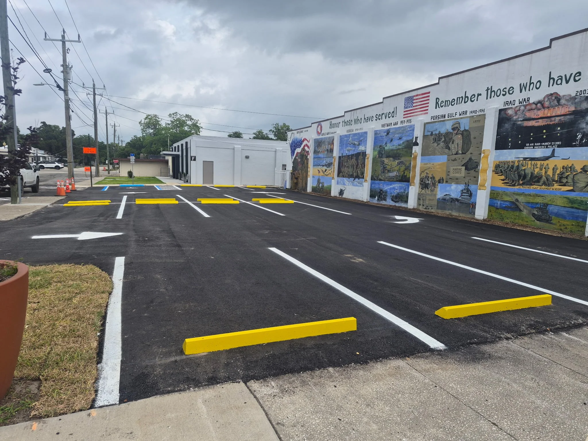 Freshly paved and painted parking lot with bright yellow wheel stops, a small white building with black stripes, and a large mural on the wall honoring military service, under a cloudy sky.