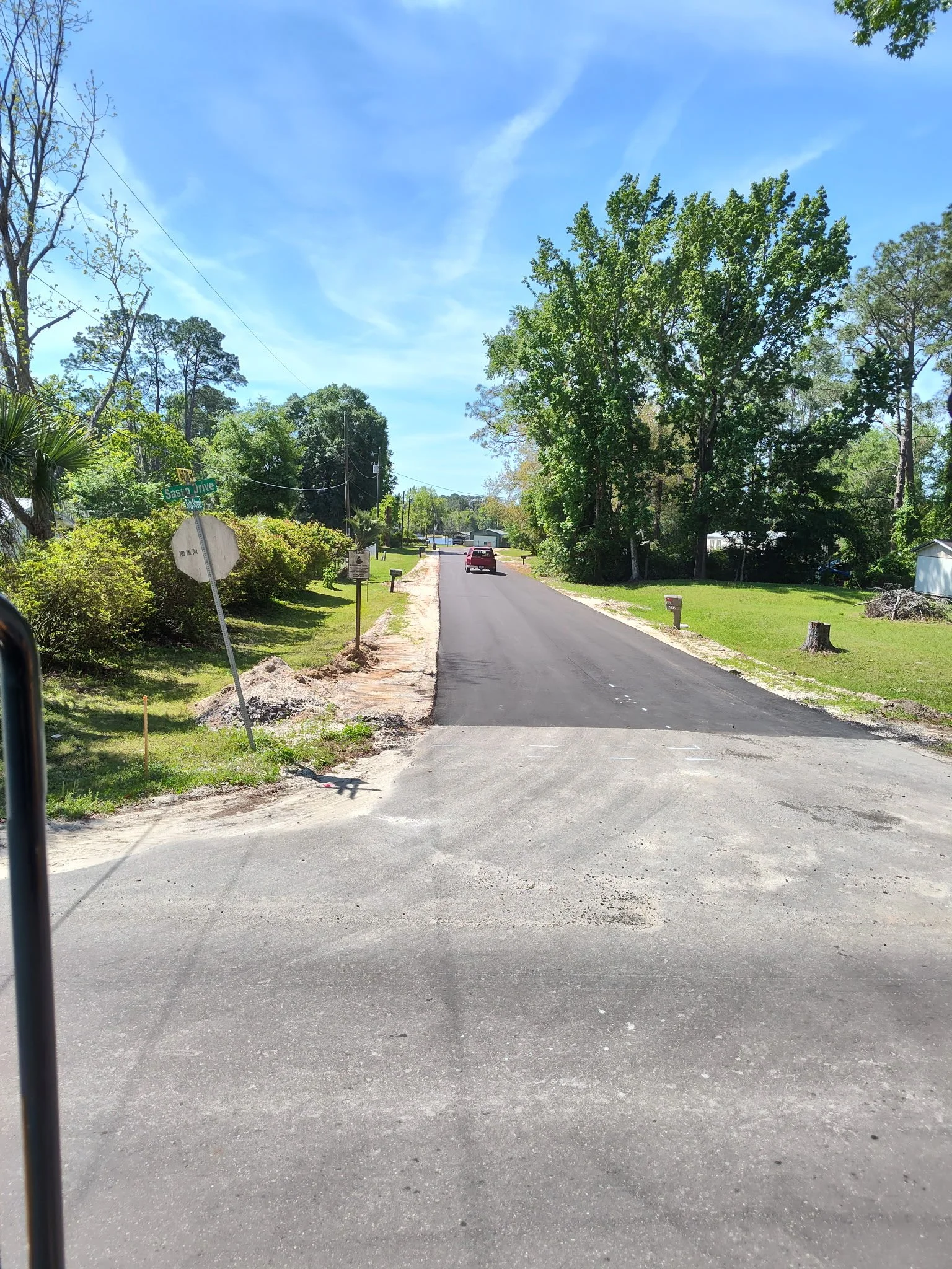 View of a residential street with trees, a parked red car, and new asphalt on the road. Signs include a street sign labeled 'Sasso Drive' and a no parking sign. The sky is clear with some wispy clouds.