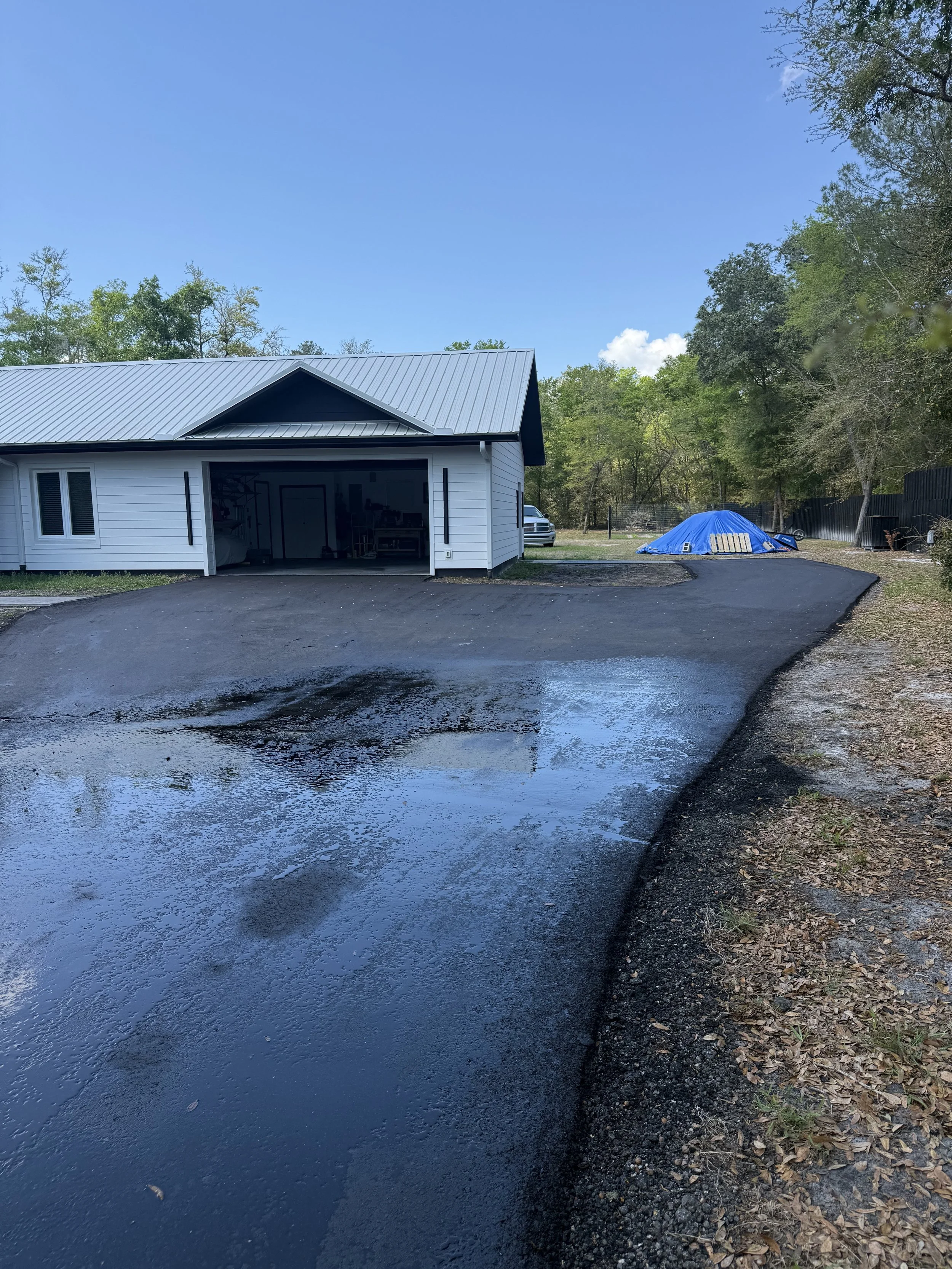 Newly paved driveway in front of a white house with an open garage, surrounded by trees under a blue sky with scattered clouds.
