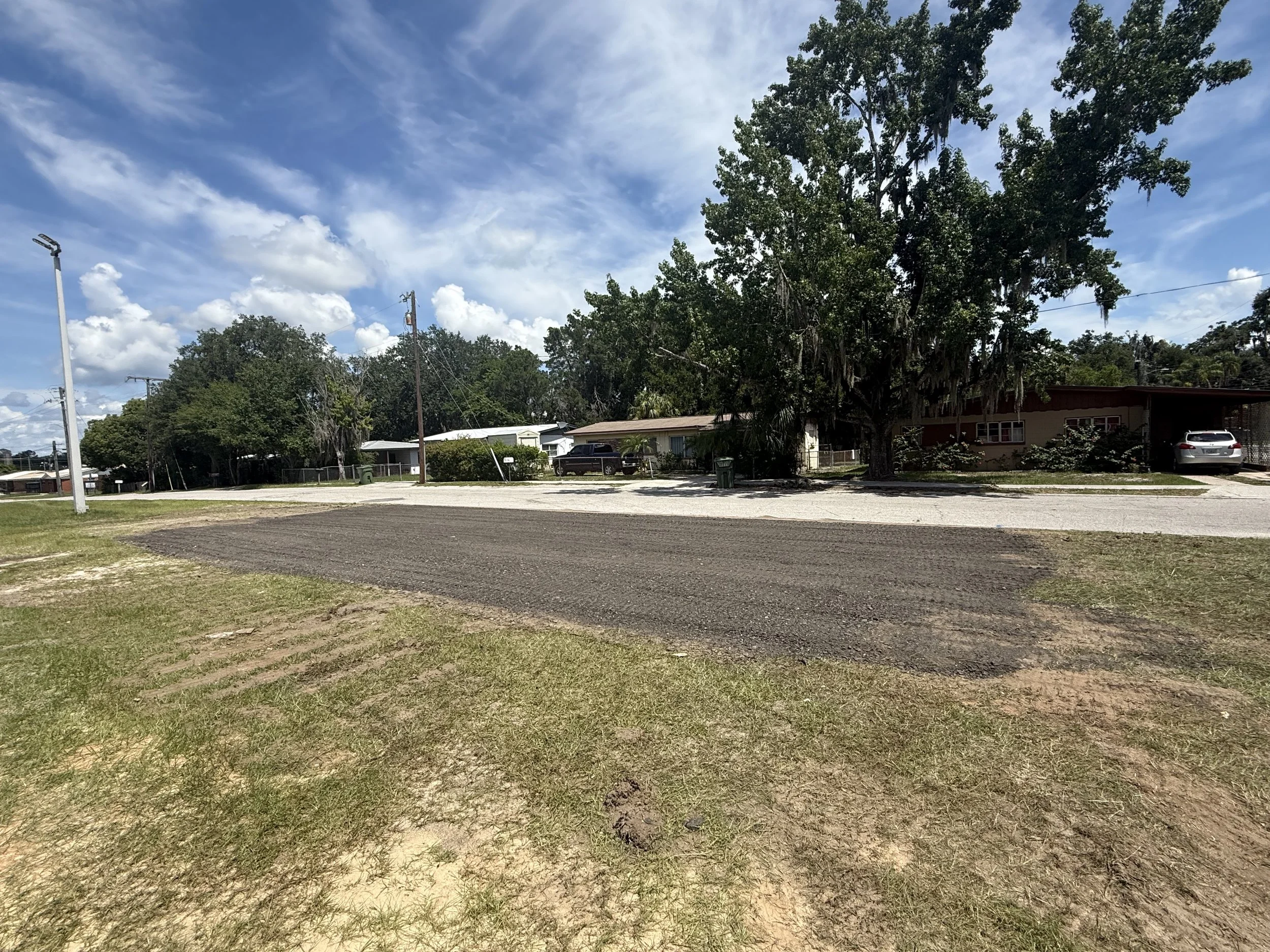 Residential neighborhood with trees, parked cars, a house, utility poles, a paved asphalt pad, and a partly cloudy sky.