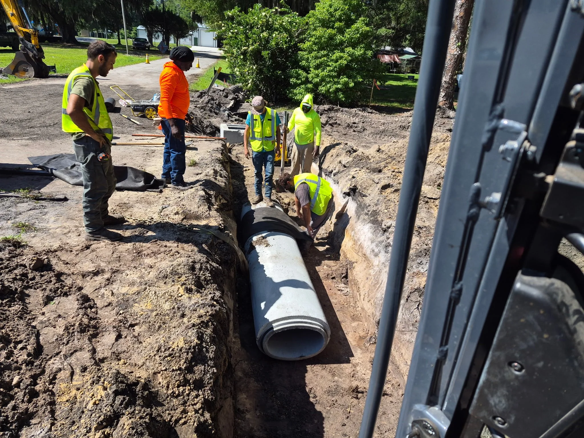 Group of construction workers working outdoors on a trench for pipe installation, with safety vests and equipment, surrounded by dirt and greenery.
