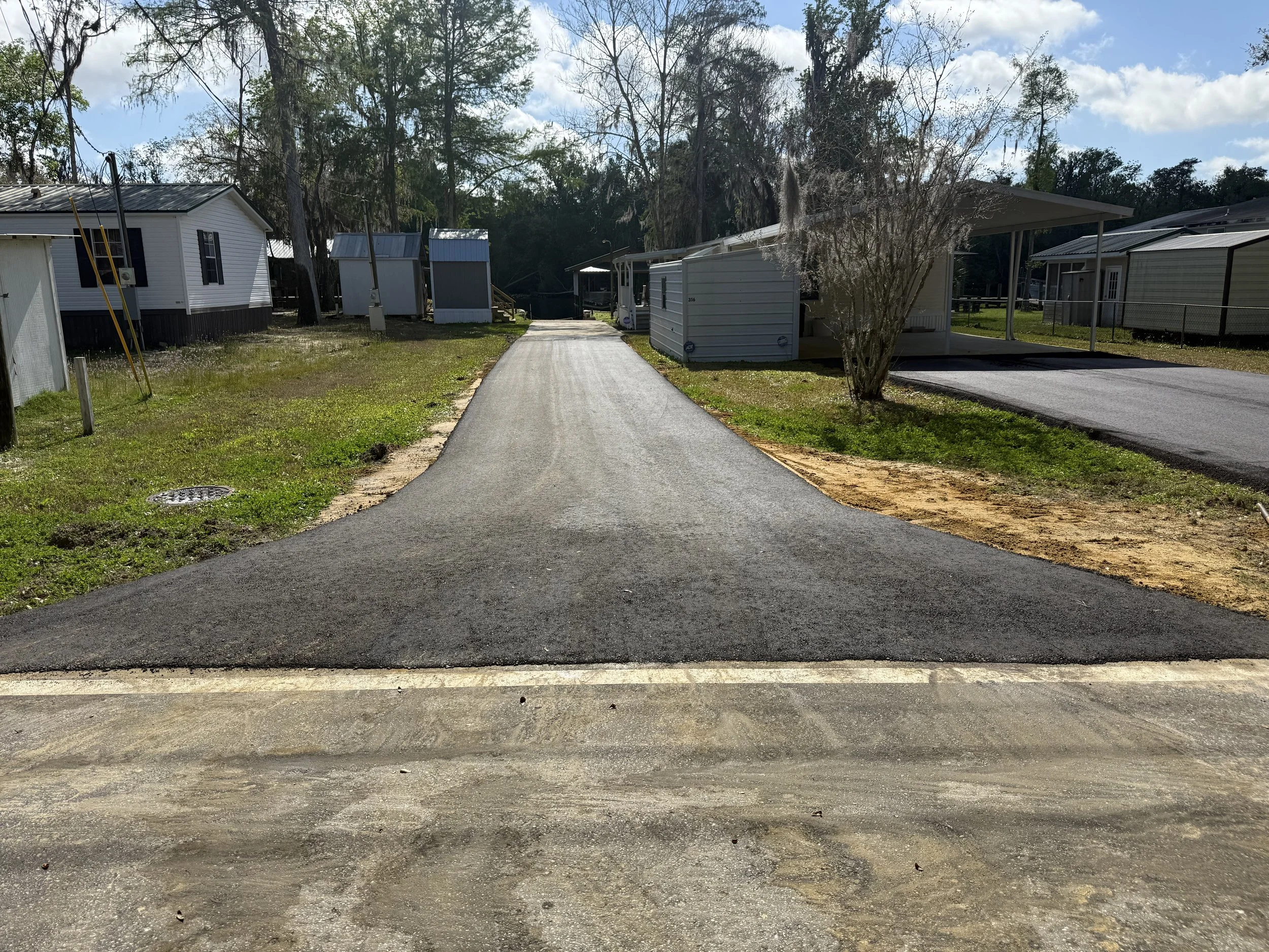 A paved driveway leading through trailers and mobile homes, with trees and a partly cloudy sky in the background.