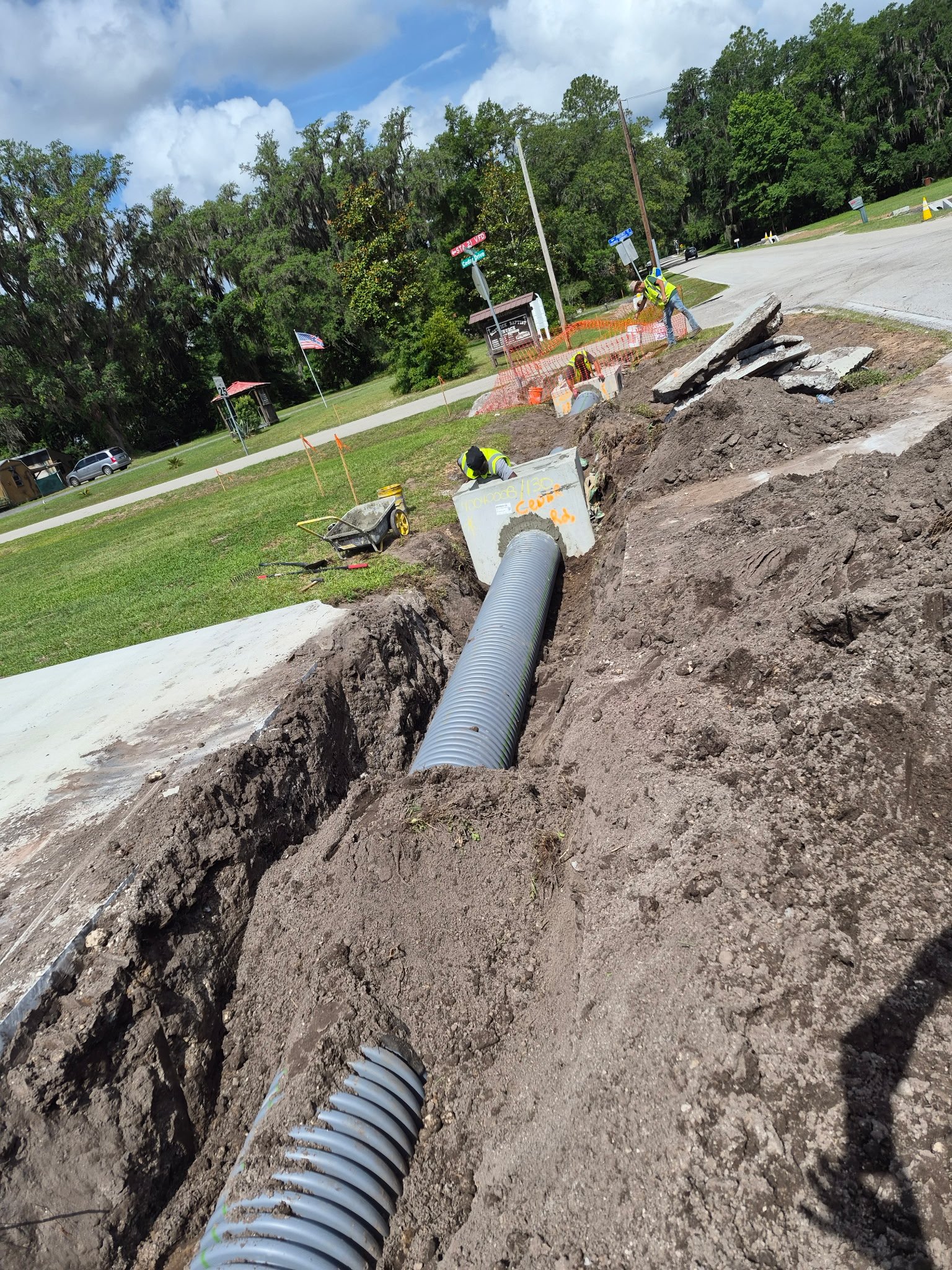 Workers installing underground drainage pipe in a trench along a roadside on a sunny day with some clouds. Orange safety fencing surrounds the work area, and construction signs are visible. Trees and parked cars are in the background.