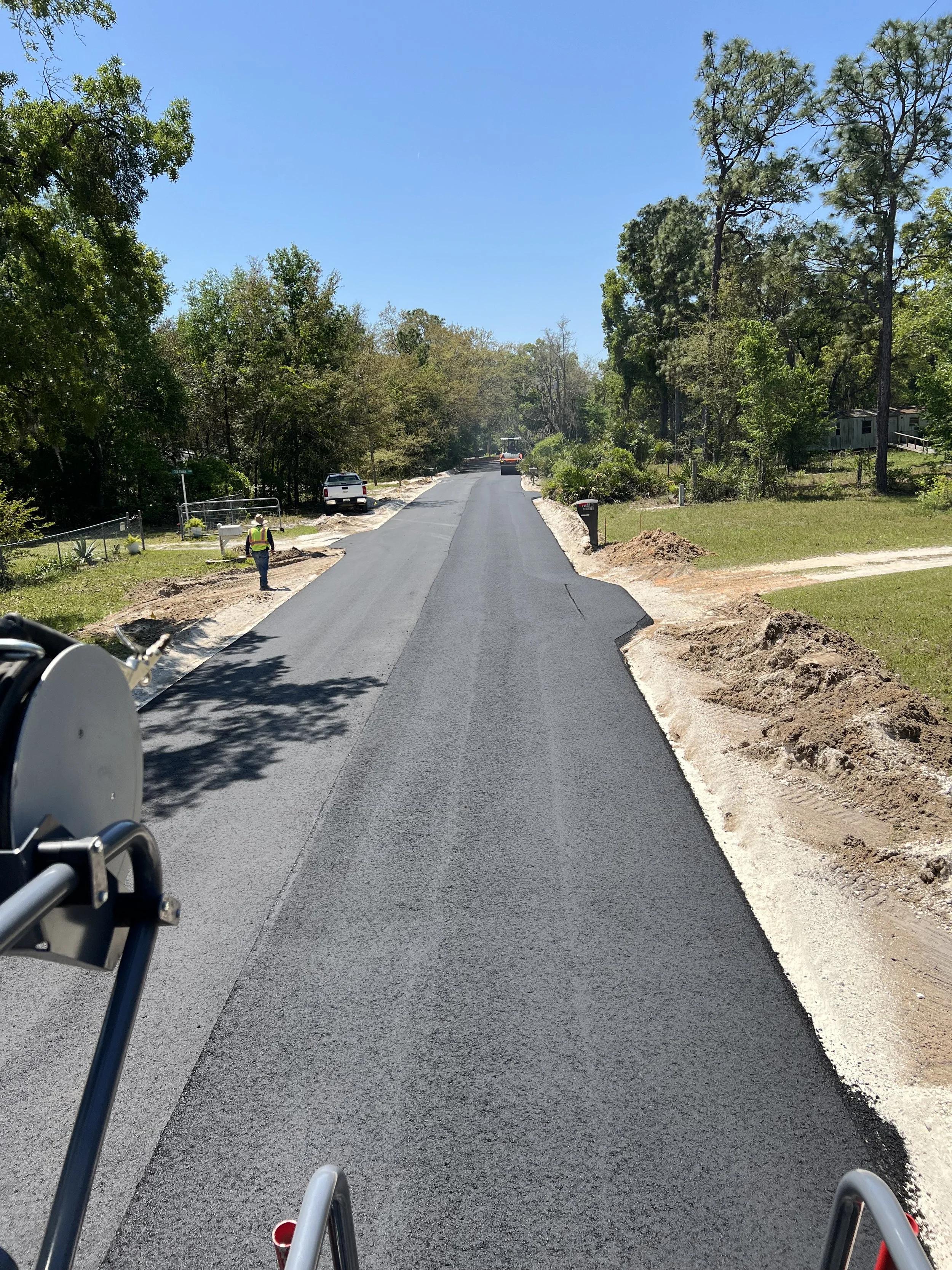 A newly paved road with construction work ongoing on the sides, a construction worker in a yellow safety vest and hat stands on the left side of the road, and a white pickup truck is parked nearby. Trees and a small house are visible in the backgroun