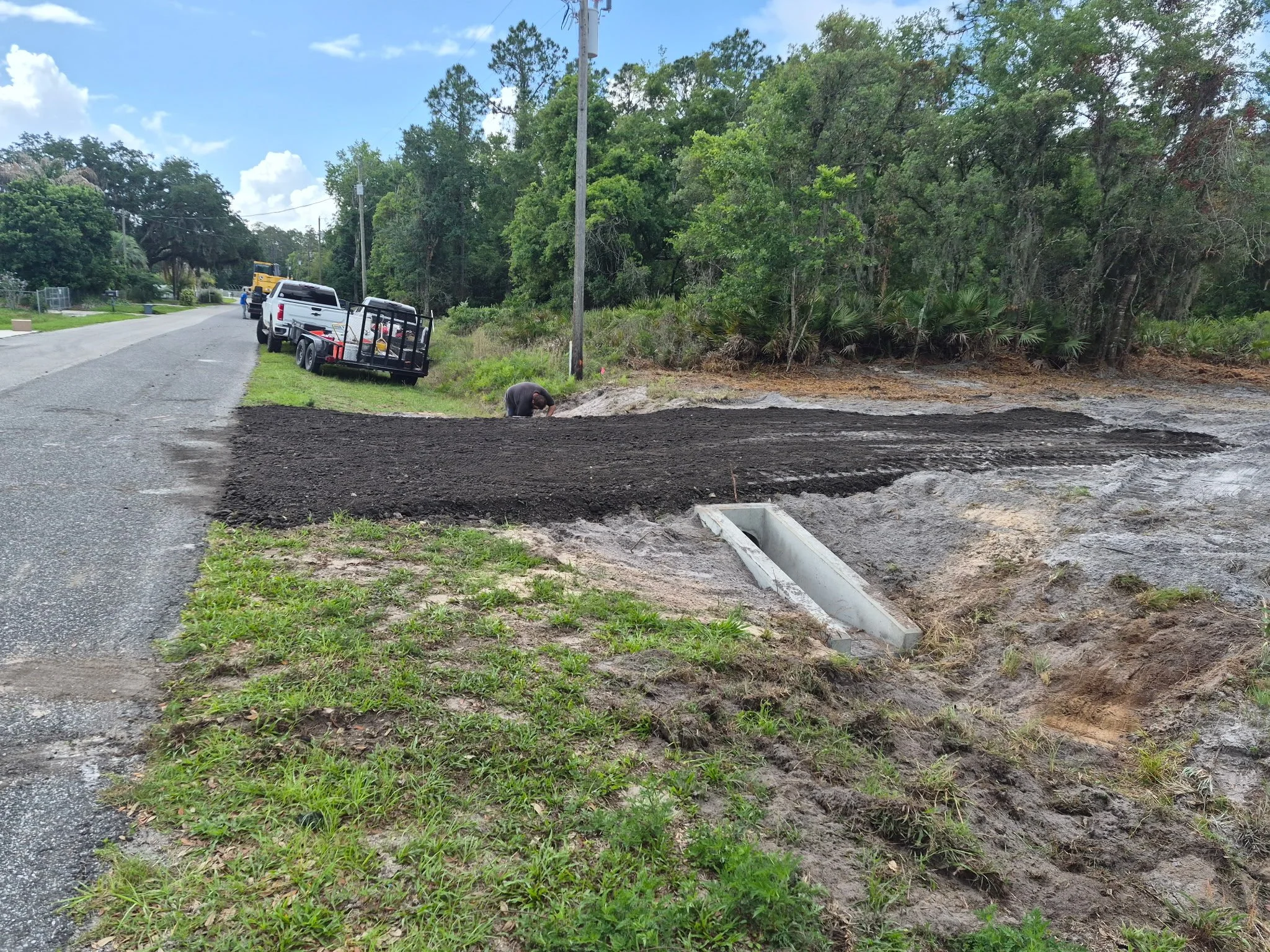 A person working on am asphalt millings apron to a dirt driveway next to a paved road, with trees and bushes in the background and a parked trailer with tools nearby.