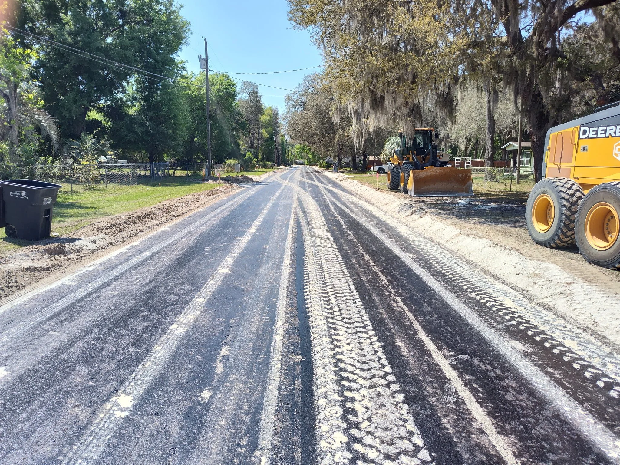 A street under construction with black asphalt patches, surrounded by trees and houses, and construction equipment on the side.