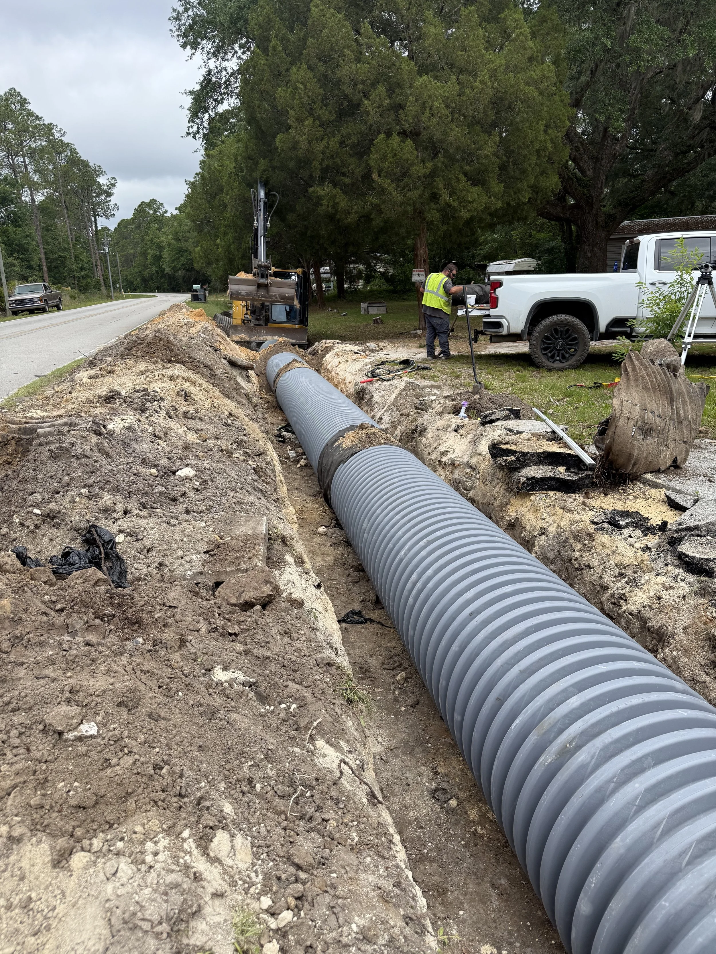 Workers installing underground drainage pipe along the side of a road, with construction equipment and a pickup truck nearby.