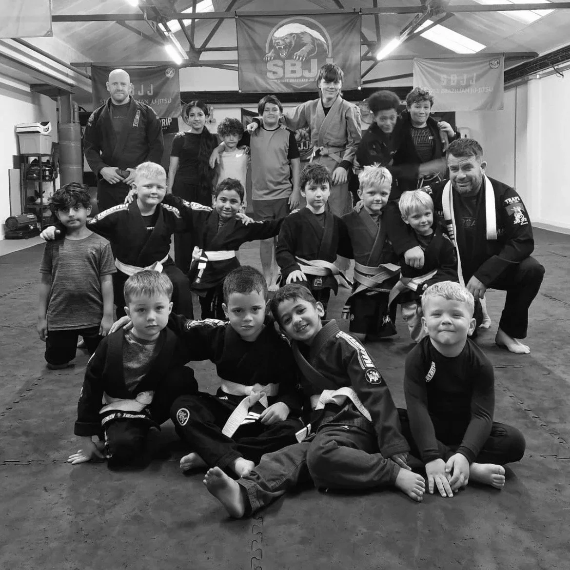 Group of children and instructors in a martial arts gym, some in jiu-jitsu uniforms and belts, posing for a photo in a dojo. The background features banners with the SBJ logo and a bear mascot.