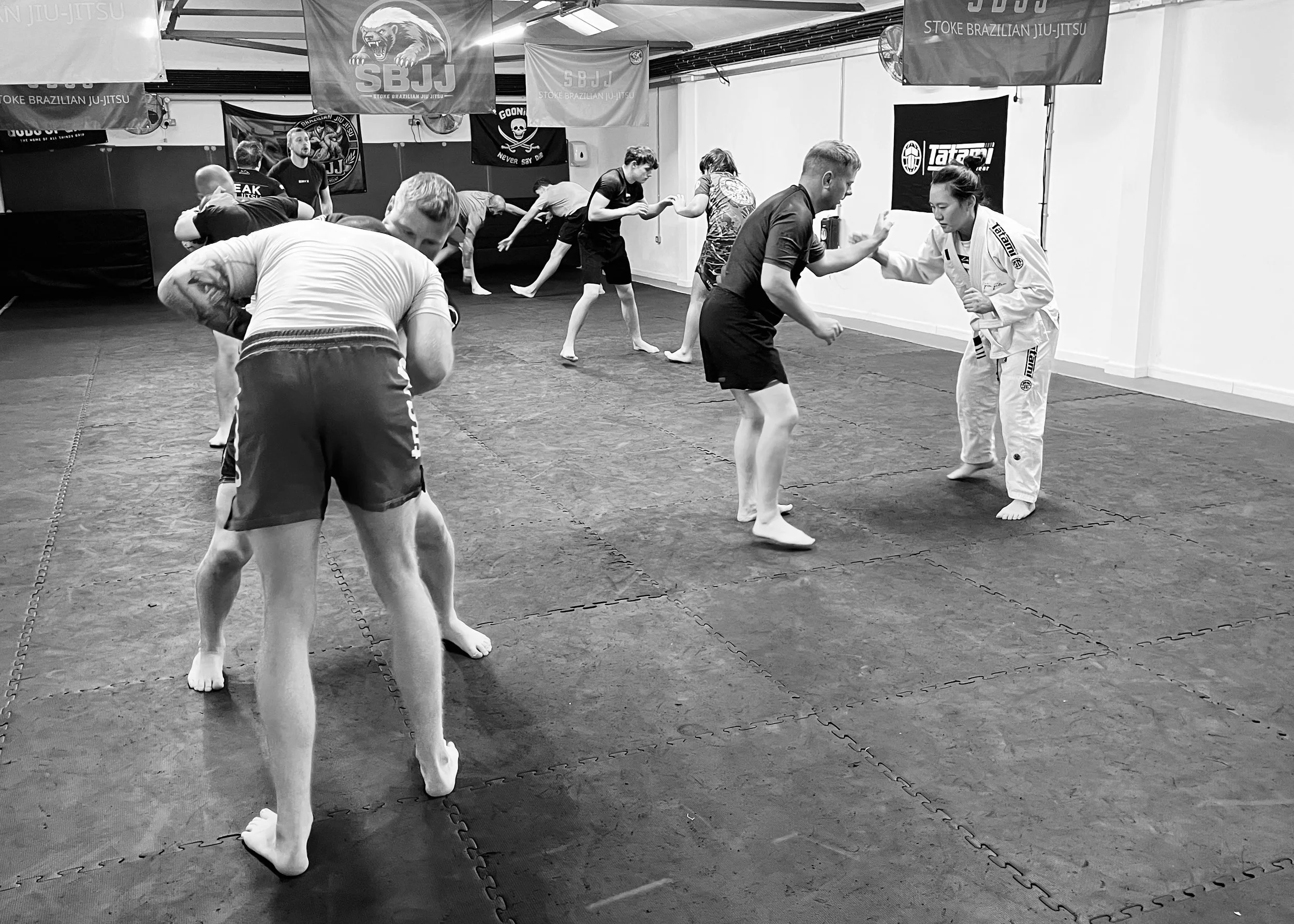 People practicing Brazilian Jiu-Jitsu in a training gym, some wearing gi uniforms, some in athletic clothing, engaging in drills and sparring on padded mats.