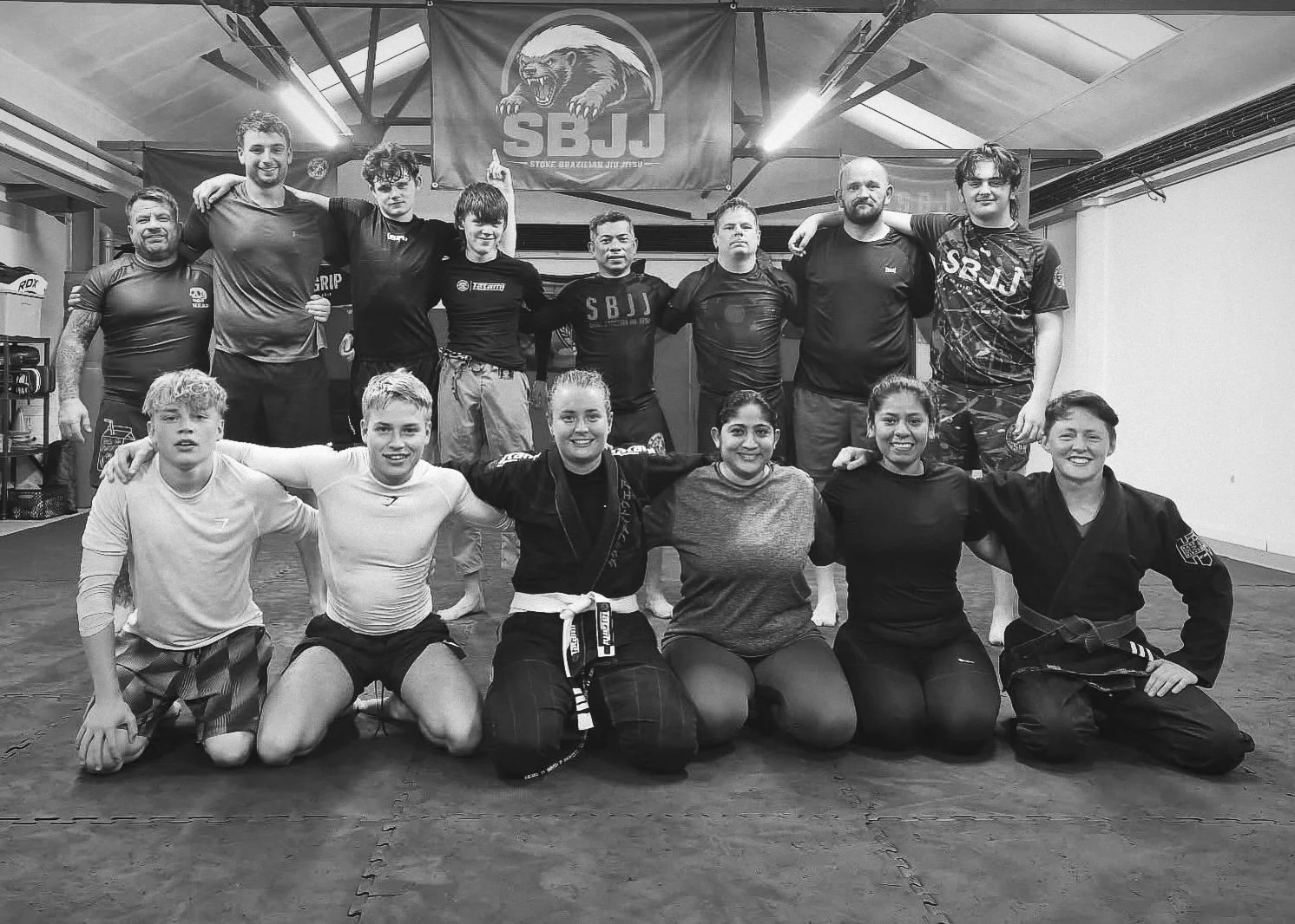 Group of martial artists, including men and women, posing together in a gym with mats on the floor, some kneeling and others standing, with a martial arts banner in the background.