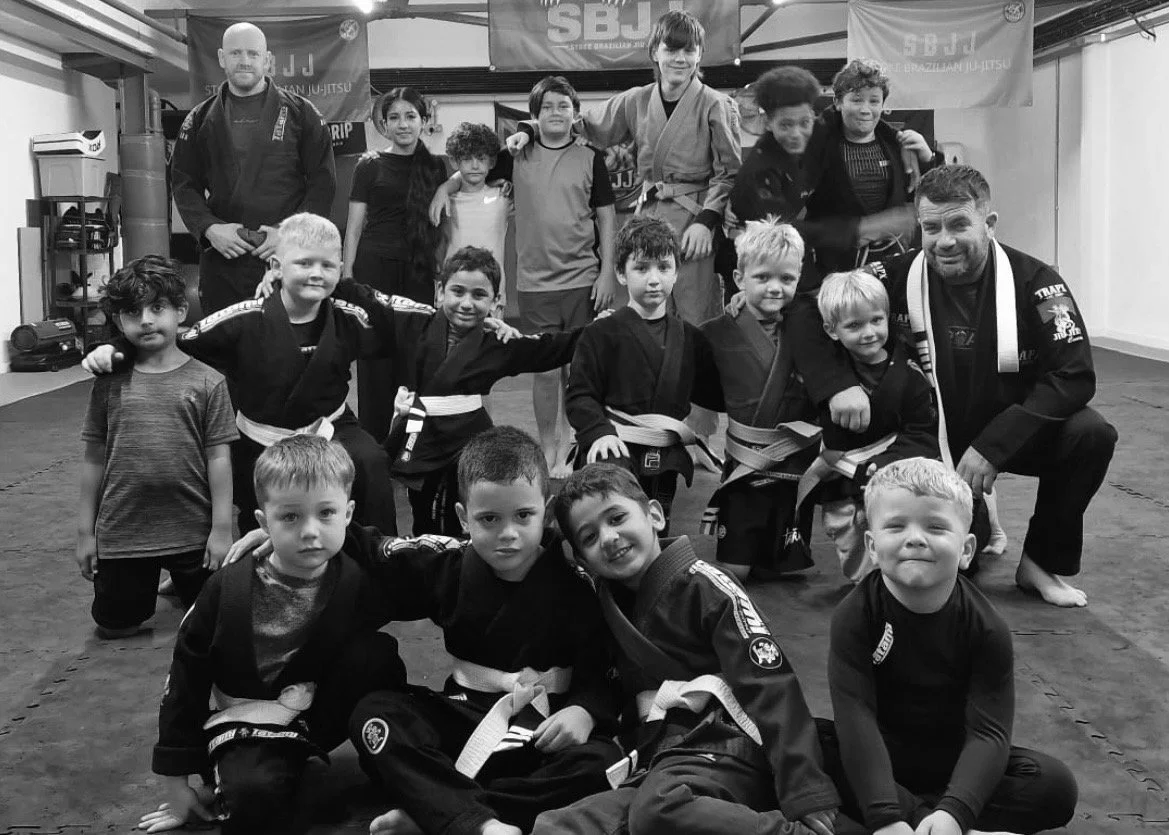 Group of children and instructors in Brazilian Jiu-Jitsu uniforms, posing in a martial arts gym.