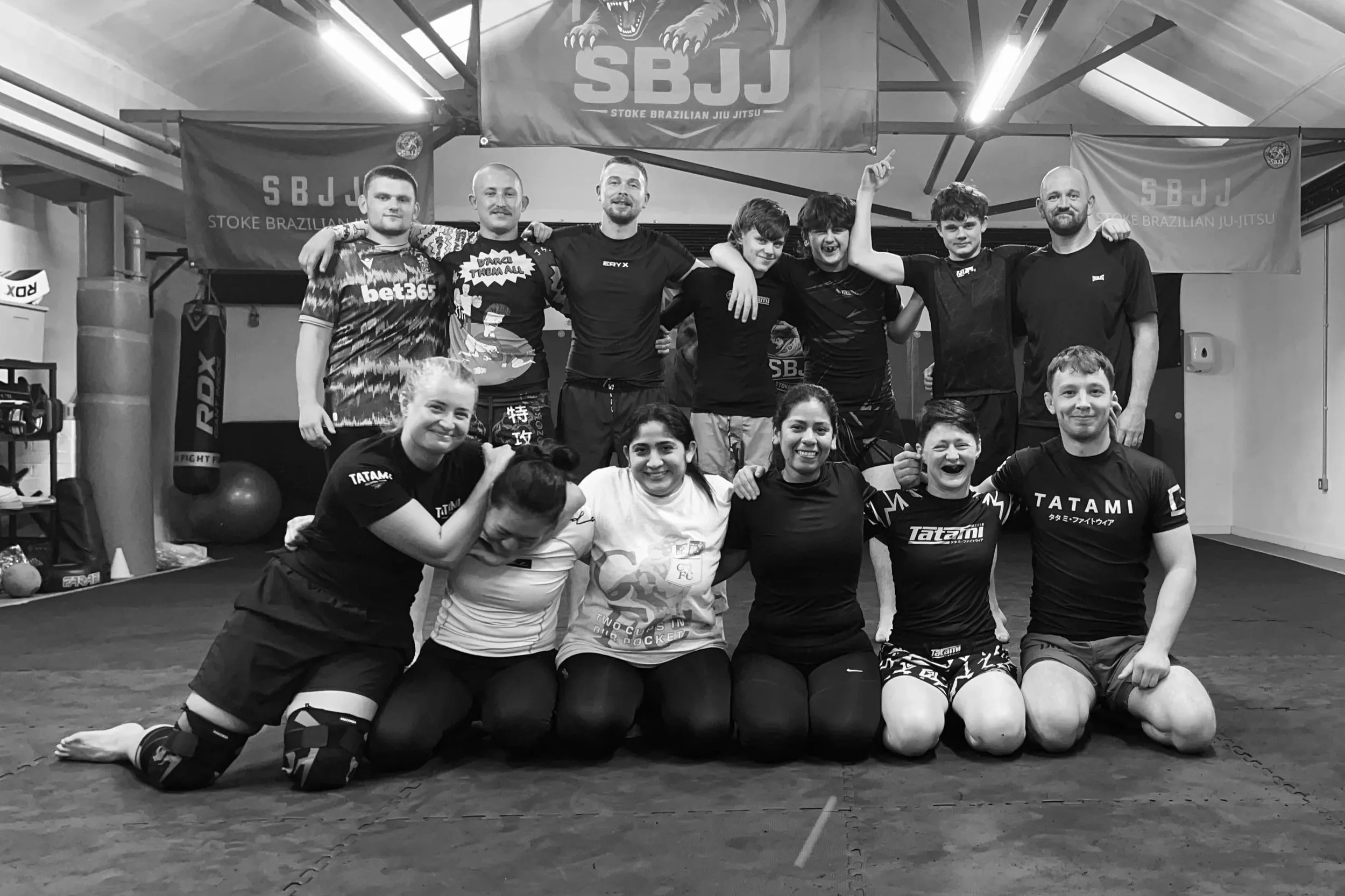 Group of martial artists posing together in a gym, some kneeling in the front and others standing behind, with banners displaying 'SBJJ' and 'Stoke Brazilian Jiu Jitsu' in the background.