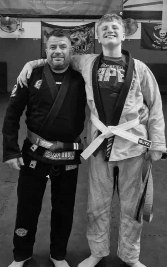 Two men in Brazilian Jiu-Jitsu uniforms smiling and posing with arms around each other in a gym, with banners on the wall in the background.