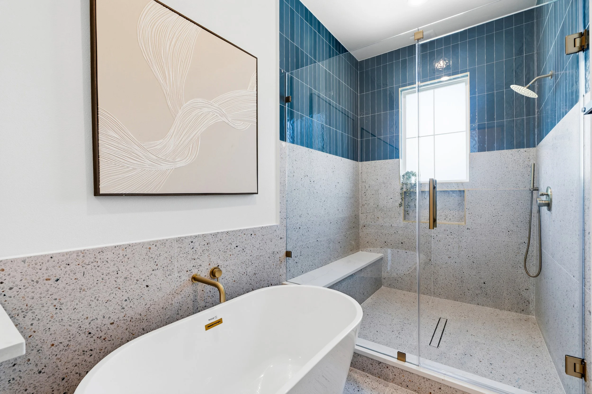 Modern bathroom with a freestanding bathtub, terrazzo walls, a glass shower enclosure with blue tiled walls, and a window with natural light.