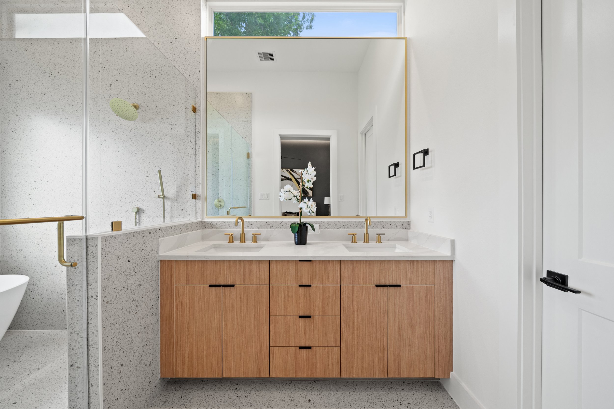 Modern bathroom vanity with a large mirror, double sink with gold fixtures, light wood cabinet, and a potted white orchid on the countertop. A shower with a glass door and speckled tile walls is visible on the left, and a window with a view of greenery and a blue sky is above.
