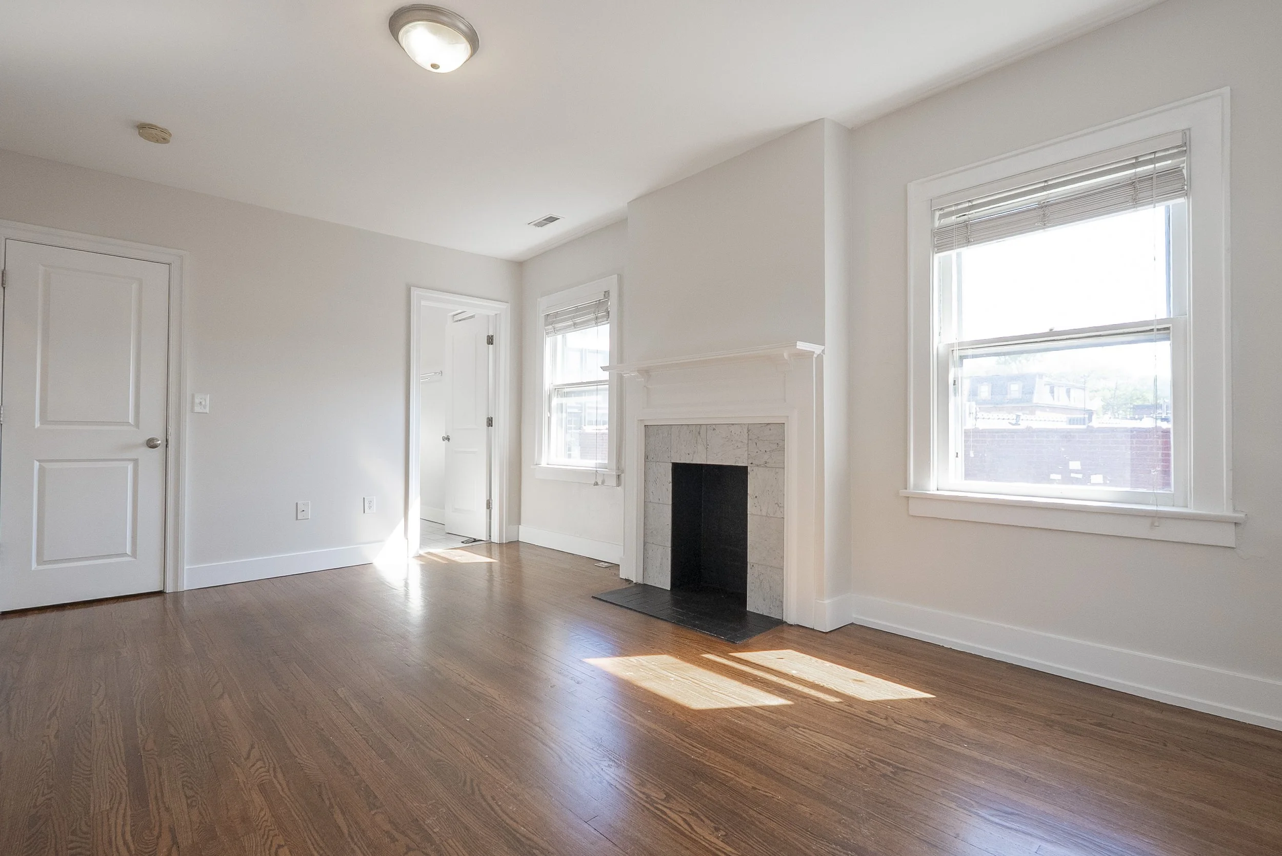 vacant real estate photos of a living room with white walls, hardwood flooring, a fireplace, three windows with blinds, a ceiling light fixture, and a door.