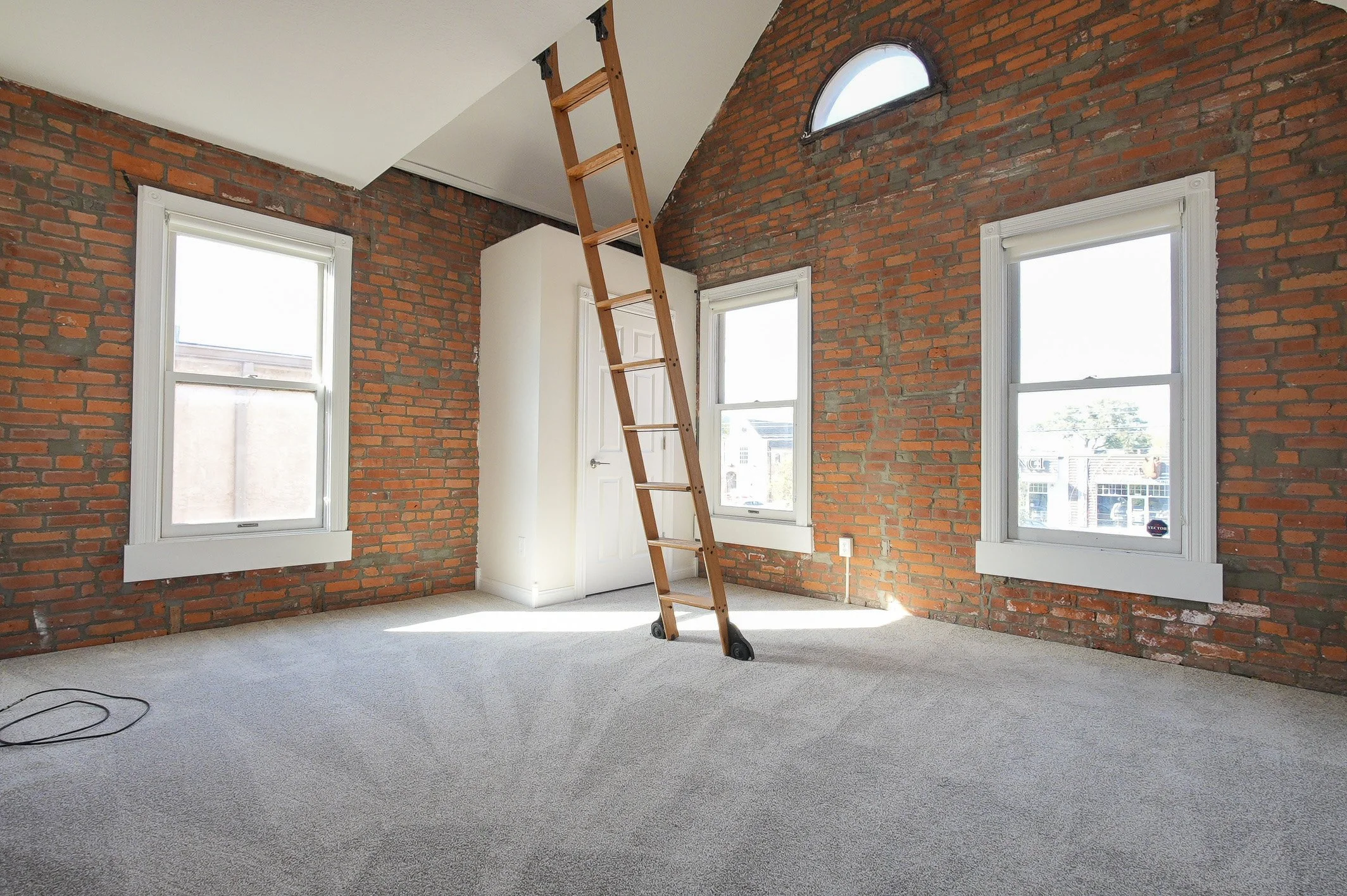 vacant listing photos with red brick walls, three large windows, a white door, a wooden ladder, and a gray carpeted floor.