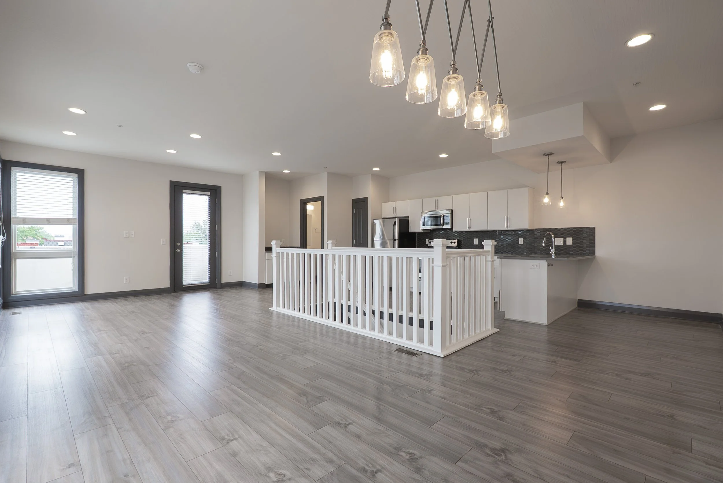 real estate photography of anInterior of a modern, open-concept apartment or house with white walls, gray trim, wood-look flooring, a white railing, and kitchen appliances visible in the background. Ceiling lights are turned on.