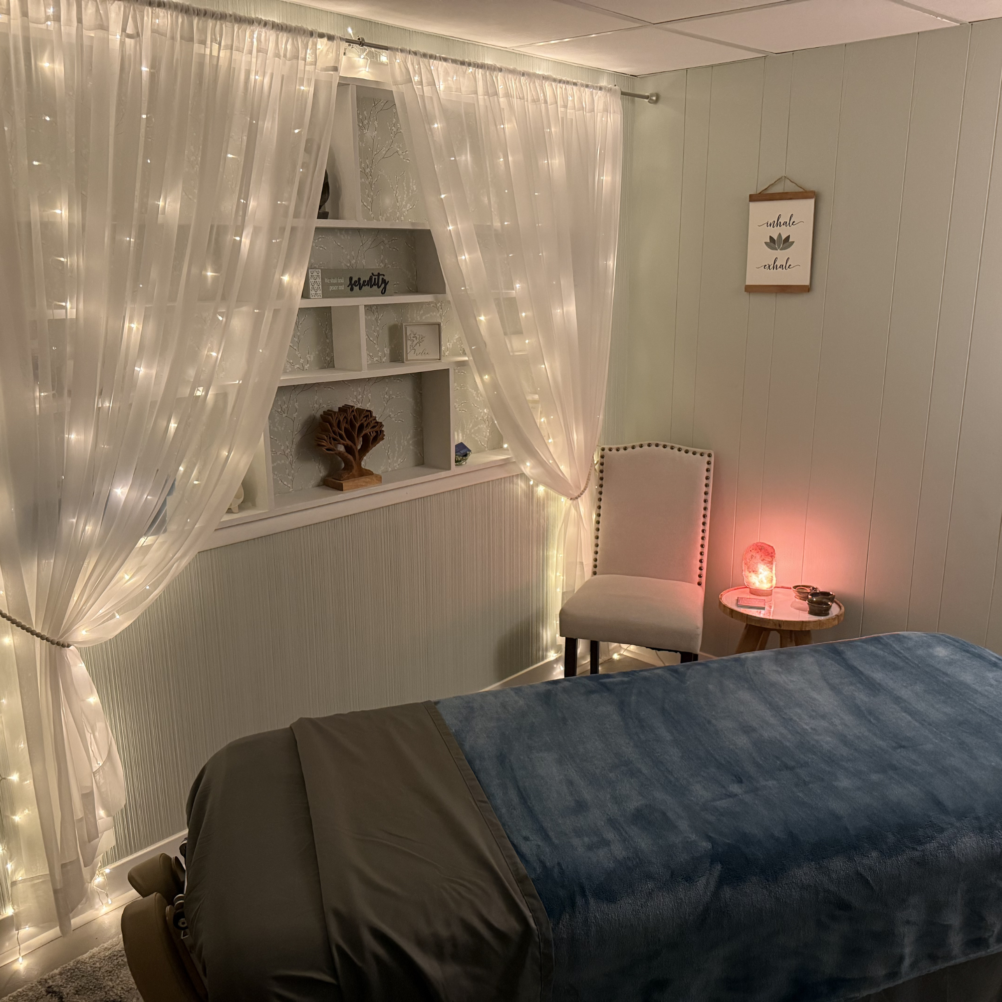Cozy bedroom with sheer curtains lit by string lights, a bookshelf with decorative items, a small table with a salt lamp, and a sign on the wall reading 'inhale, exhale'.