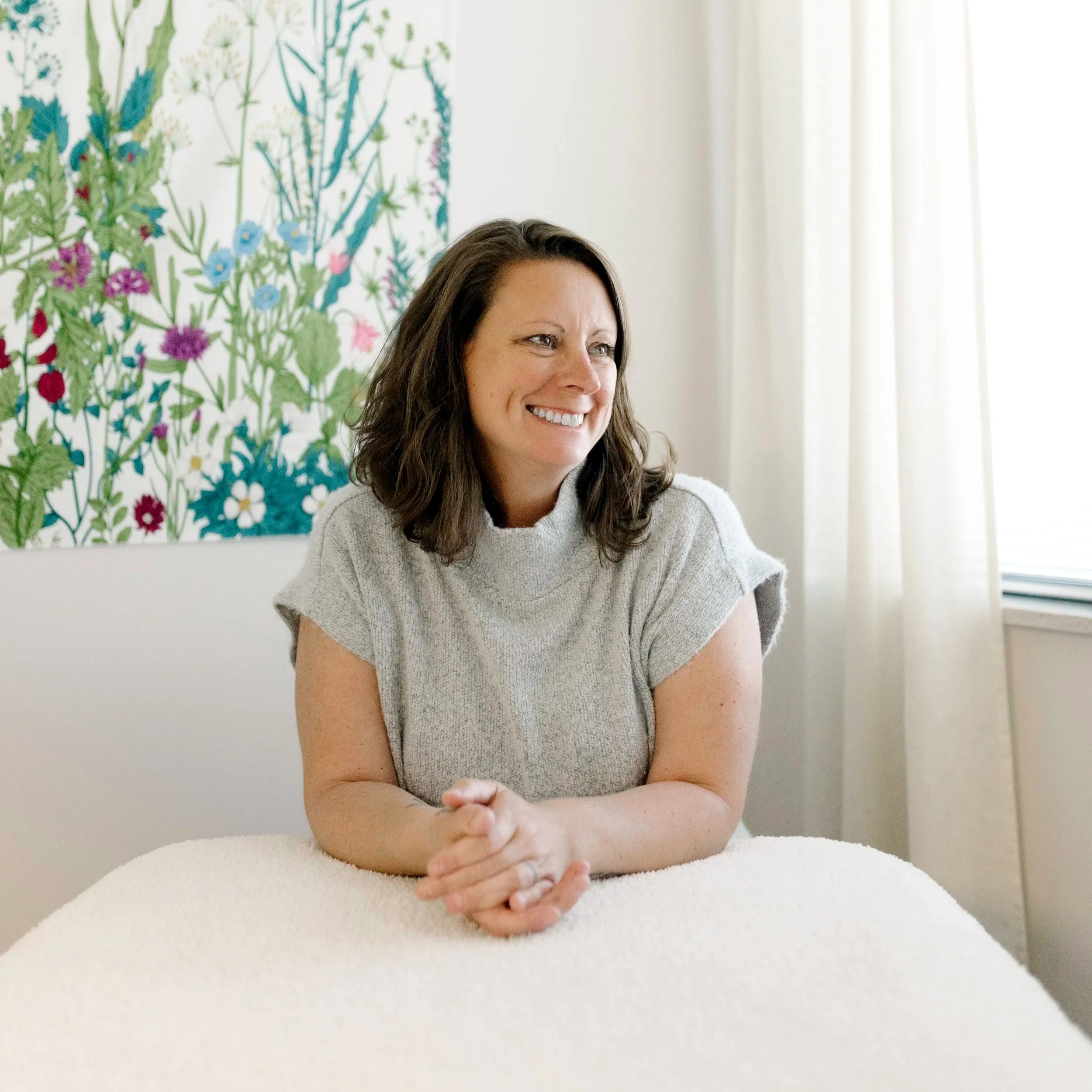 Woman sitting at a white table with her arms resting on the table, smiling and looking slightly to her left with a colorful botanical painting on the wall behind her and light-colored curtains to the right.