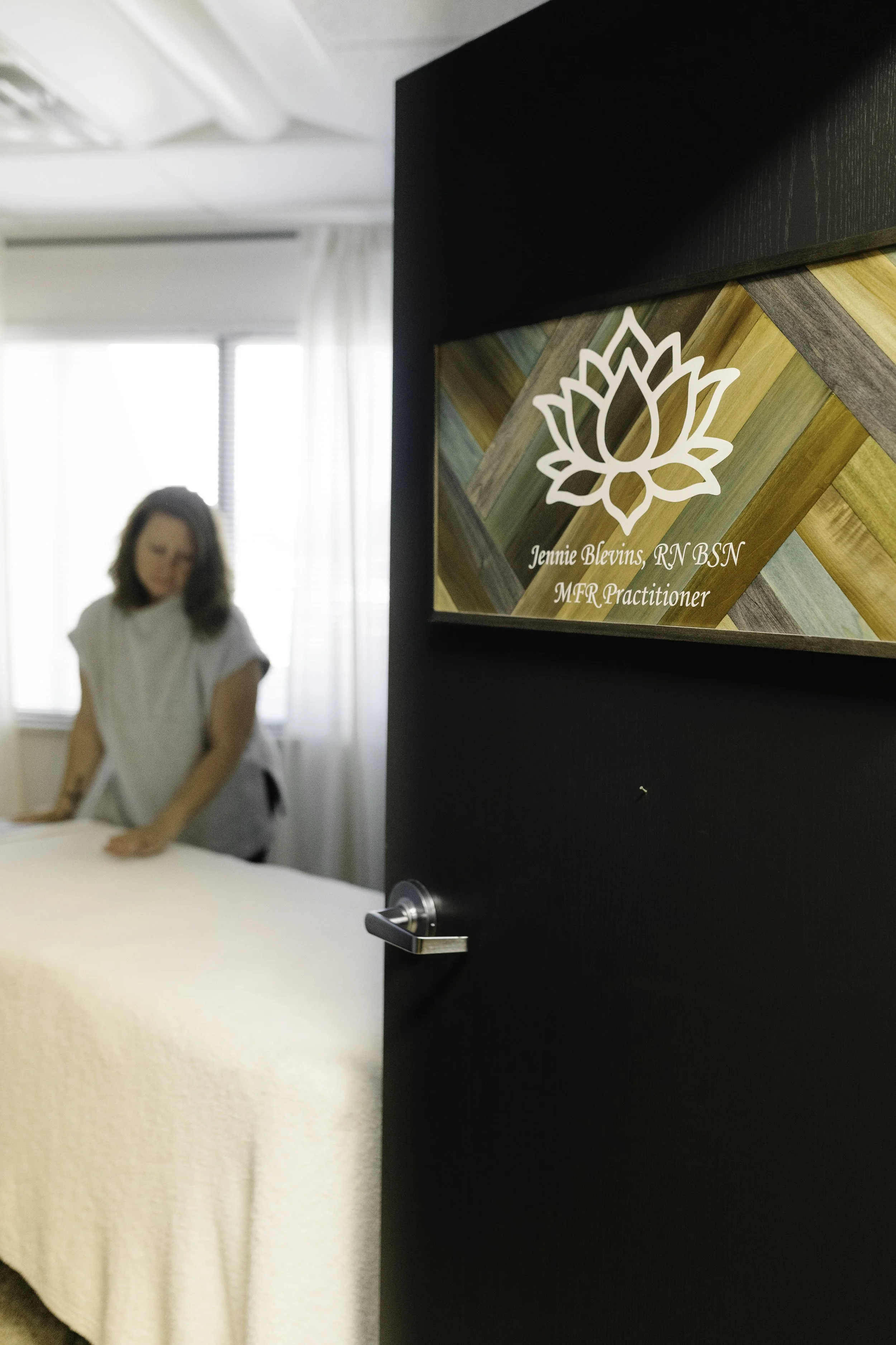 Sign inside a door with a lotus flower symbol and the name Jennie Blevins, RN BSN, MFR Practitioner, hanging on a black framed board in a bright room with white curtains and a woman in a white medical gown.