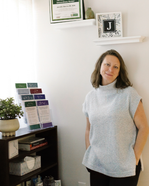 A woman standing in an office next to a bookshelf with plants and brochures, with framed documents and a decorative letter 'J' on a shelf in the background.