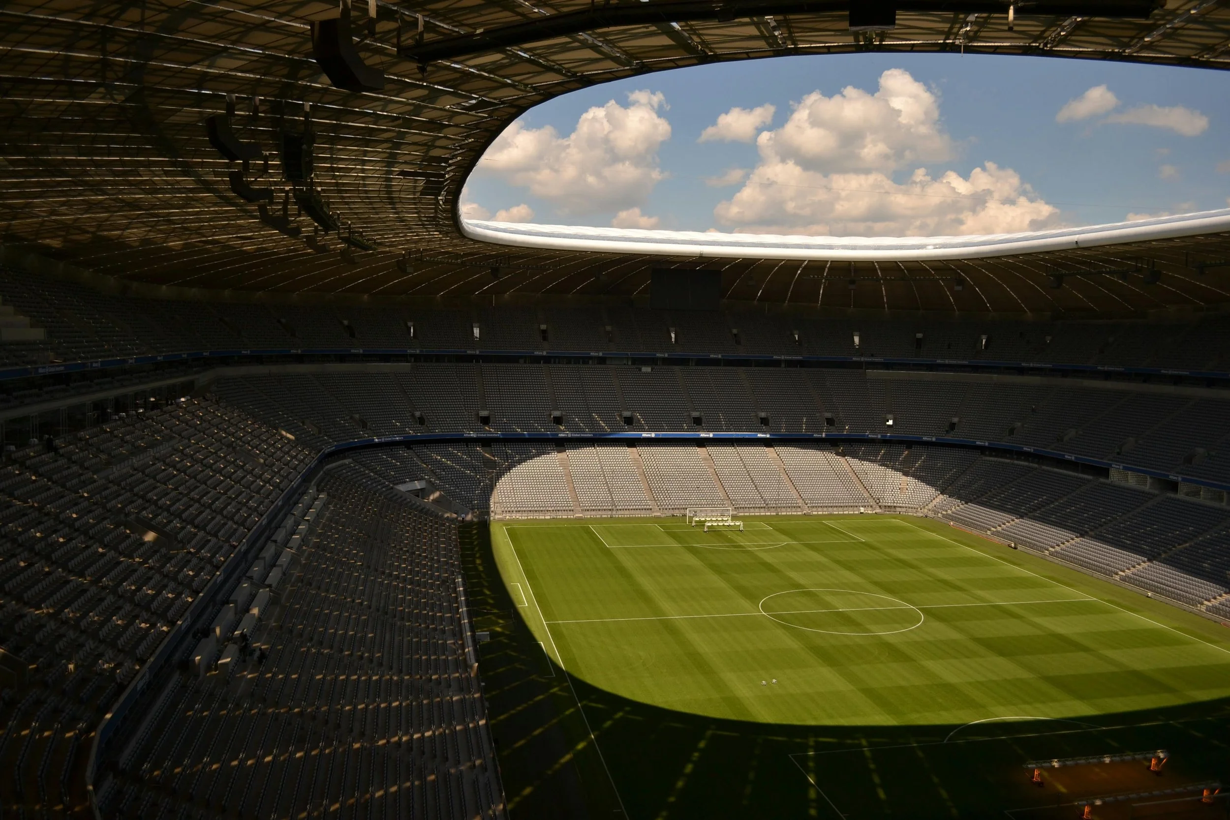 Inside a modern stadium with empty seats, a well-maintained green soccer field, and a partially open roof showing a blue sky with clouds.