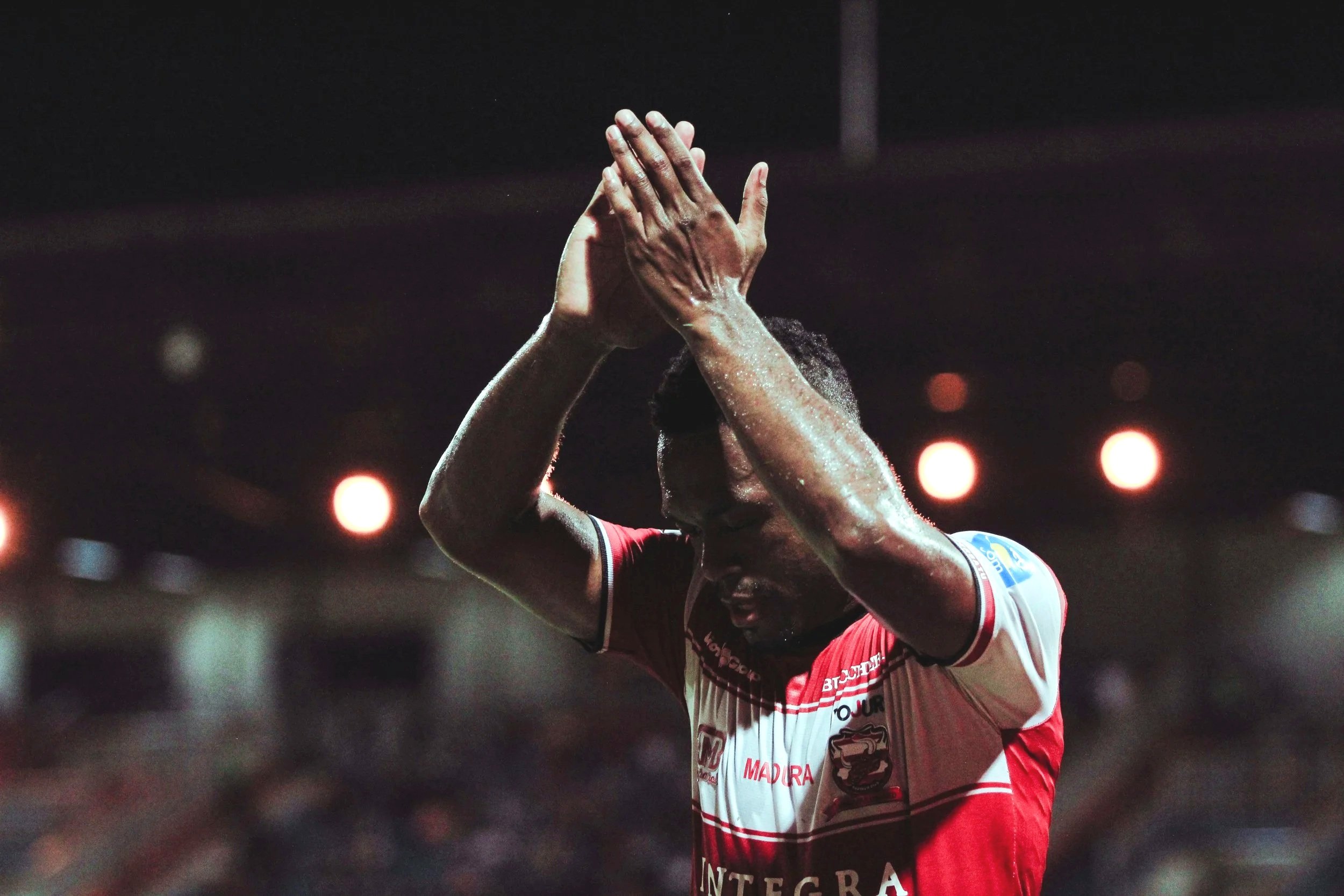 A football player in a red and white jersey with arms raised, hands clasped together, standing on a dark field with bright lights in the background.