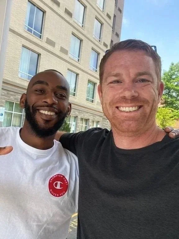 Two men smiling and taking a selfie outside in front of a multi-story building with windows.
