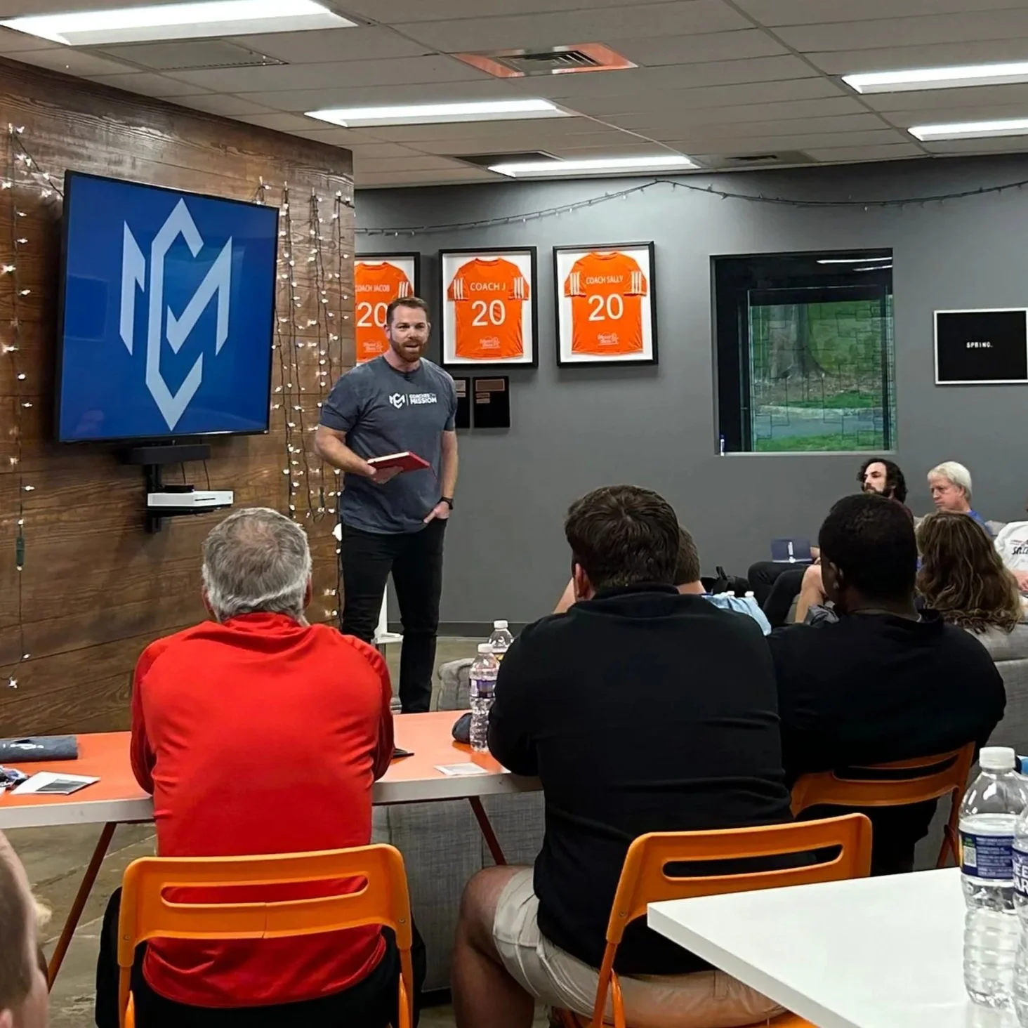 A man with a beard, wearing a gray T-shirt and black pants, is standing and speaking to an audience in a conference room. The audience is seated at tables, and some are looking at the speaker. The wall behind him has framed sports jerseys and a large blue logo on a wooden wall with string lights. There is a window on the right side, and the room is lit with ceiling lights.