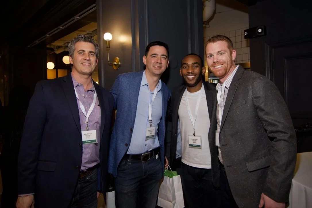 Four men wearing business attire, standing together and smiling at a professional event or conference with name tags around their necks.