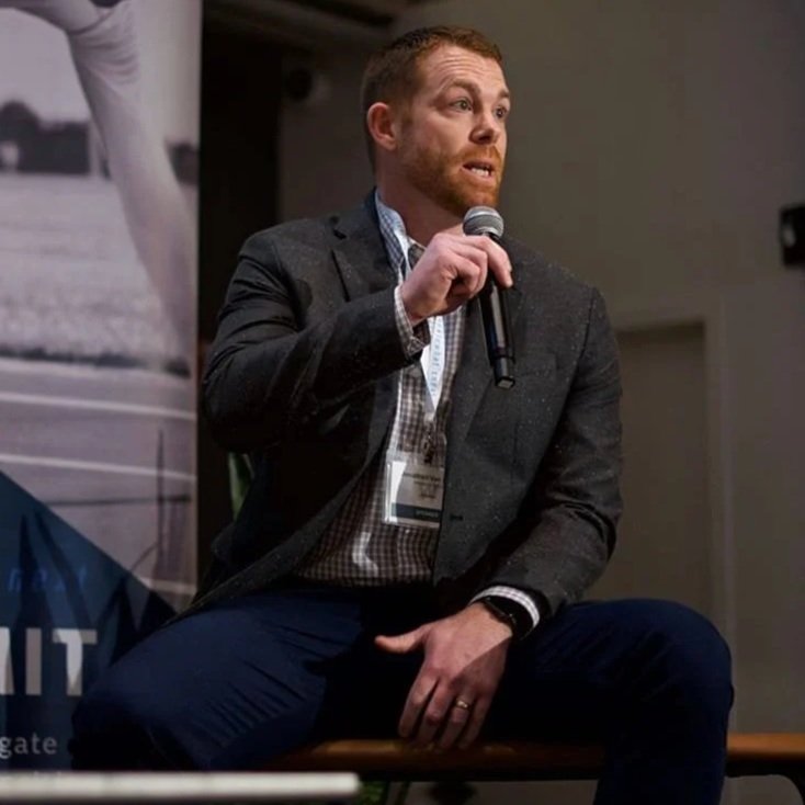A man with red hair and a beard sits on a bench, holding a microphone, speaking at an indoor event.