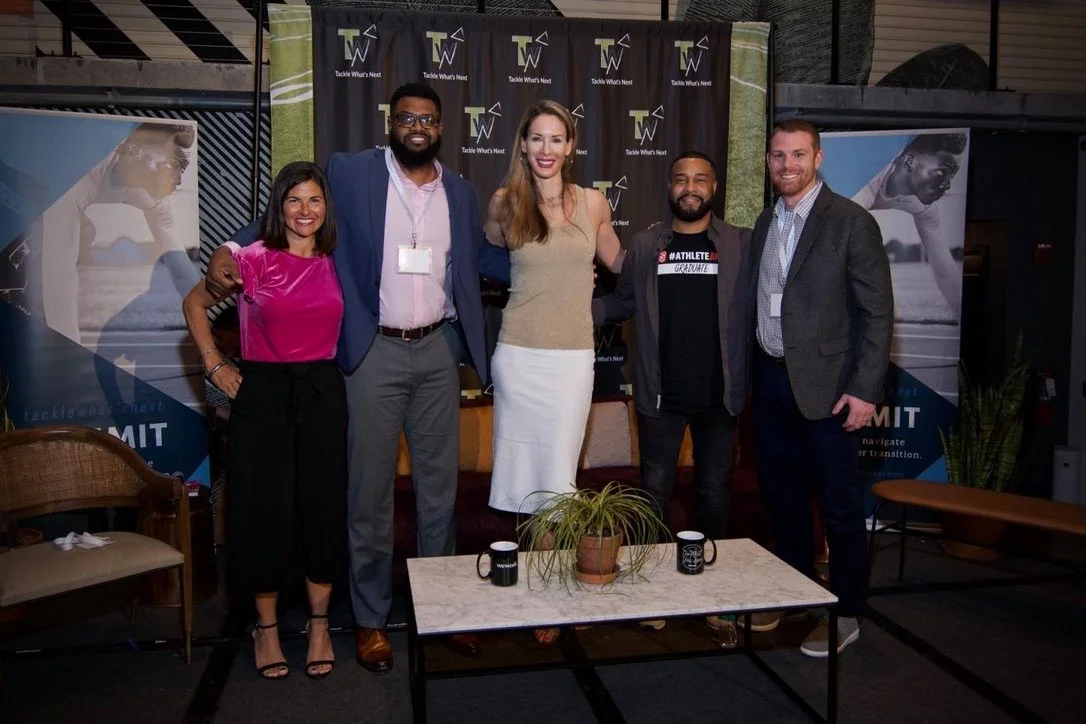 Group of five people standing together indoors at a professional event, smiling for the camera. Men and women dressed in business attire, with a backdrop and banners behind them.