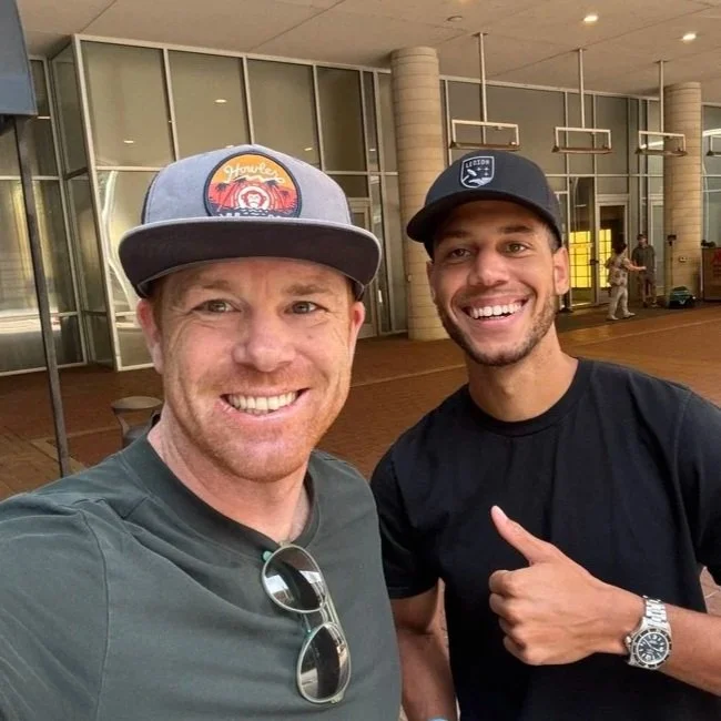 Two men smiling for a selfie in an outdoor area with glass windows and a tiled floor. The man on the left is wearing a gray cap with a colorful logo, gray T-shirt, and sunglasses hanging from his shirt. The man on the right is wearing a black cap, black T-shirt, and a silver wristwatch, and is giving a thumbs up.