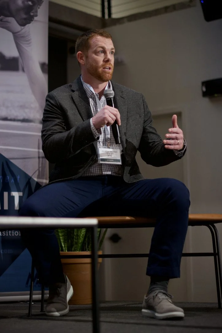 A man with red hair and beard in a suit jacket and checkered shirt, sitting on a bench, speaking into a handheld microphone during a presentation or panel discussion.