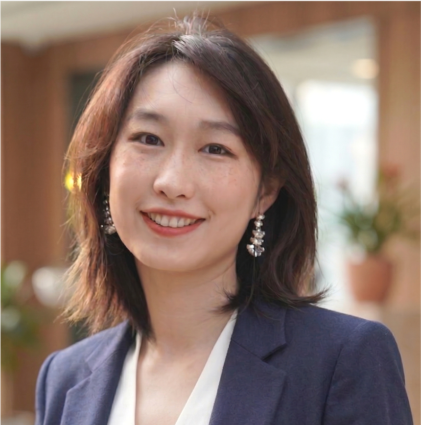 A smiling woman with shoulder-length dark hair, wearing earrings, a navy blazer, and a white top, in a well-lit indoor setting with plants in the background.