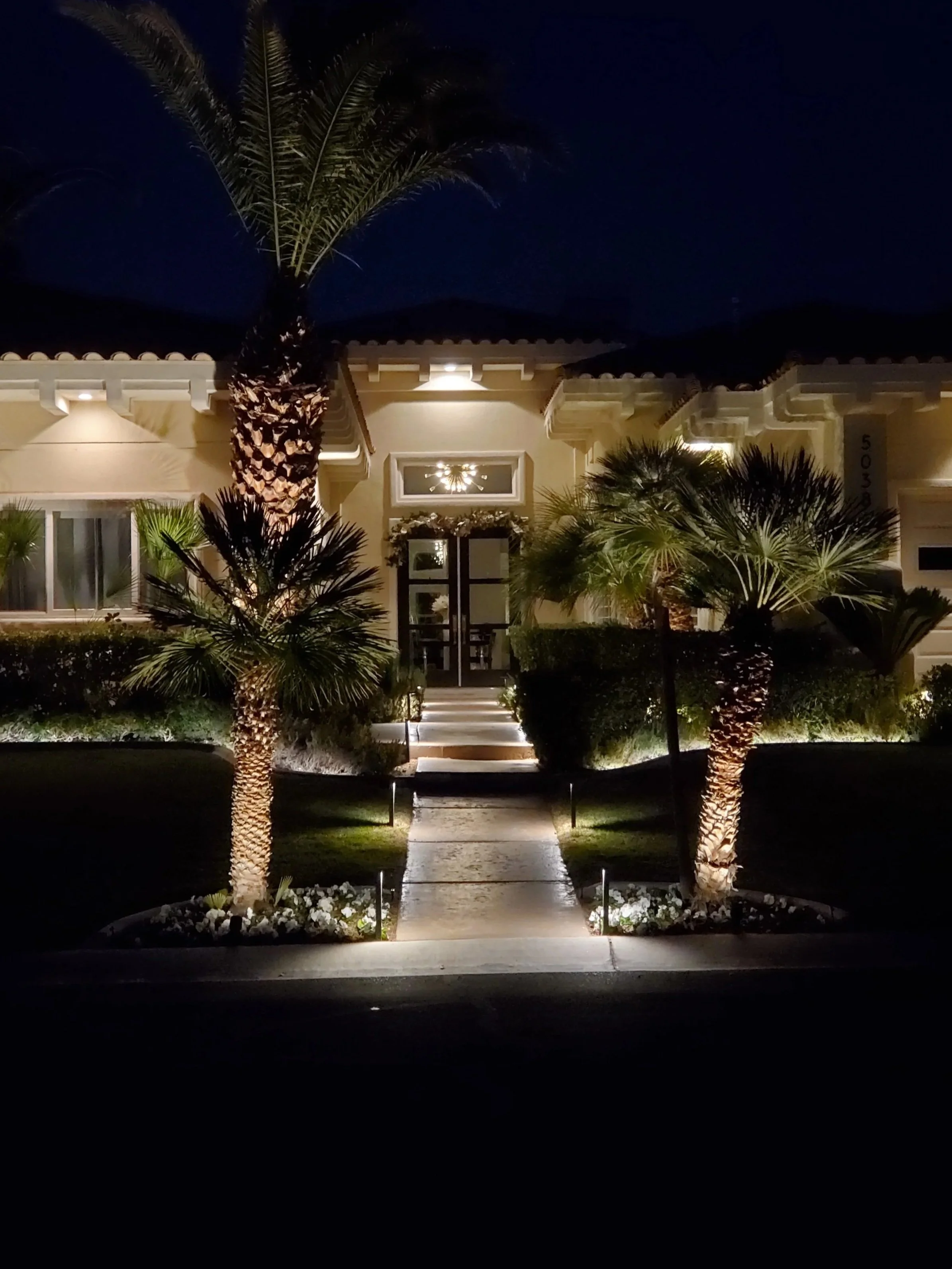 Night view of a house's front entrance with a pathway, palm trees, and exterior lighting.