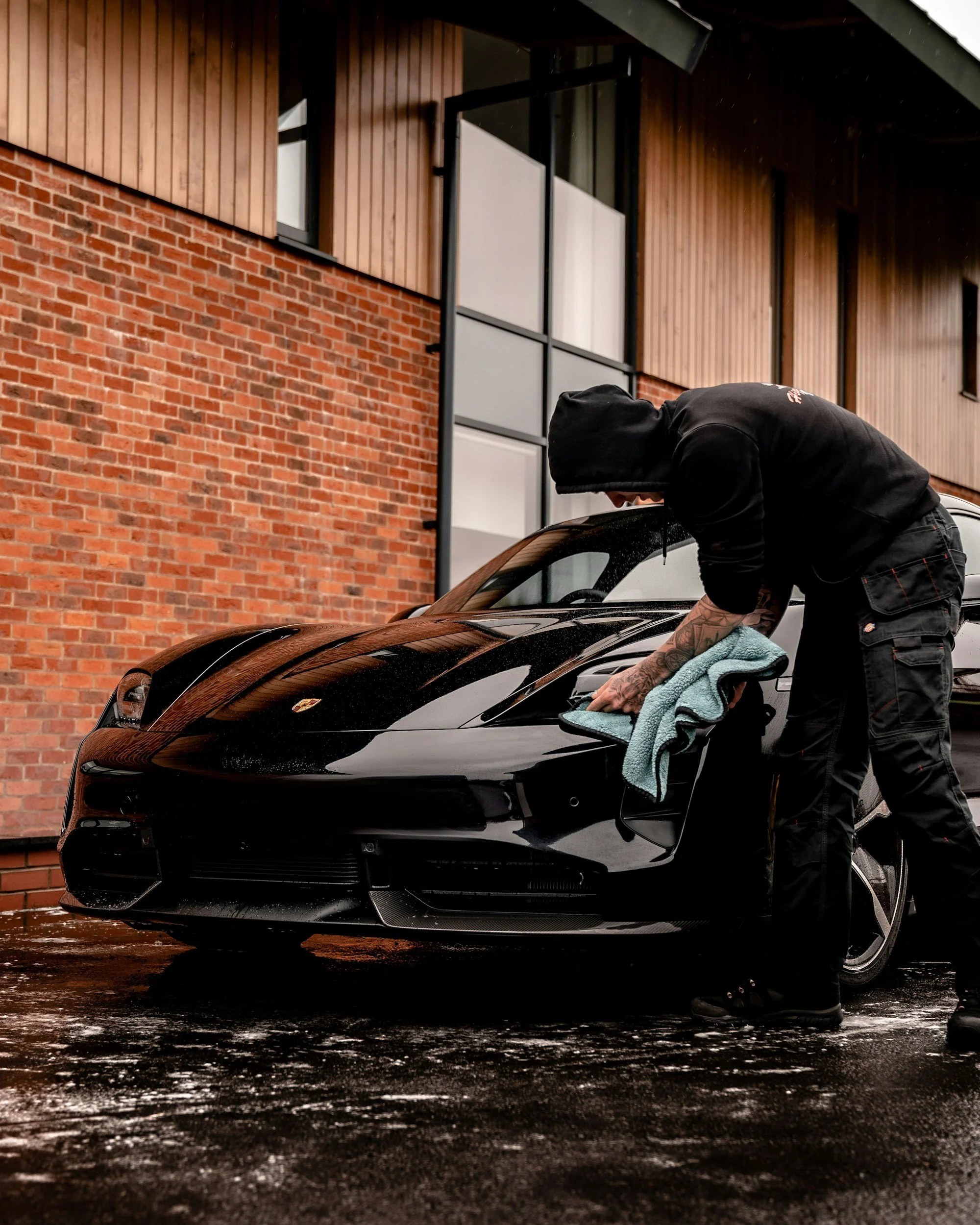 A person dressed in black with a hood and tattoos is washing a black sports car with a towel outside a brick building.
