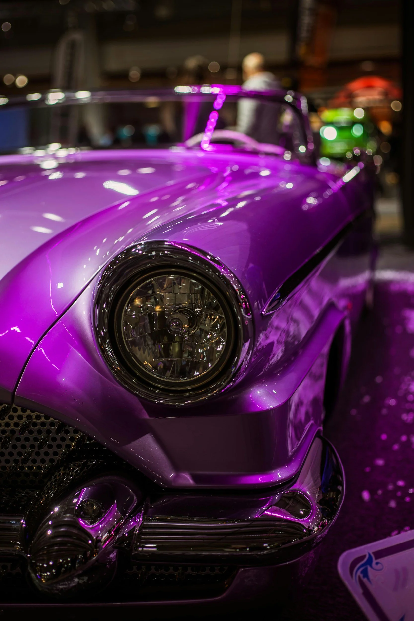 Close-up of a shiny purple sports car with a prominent headlight and sleek design, illuminated by purple and pink lighting.