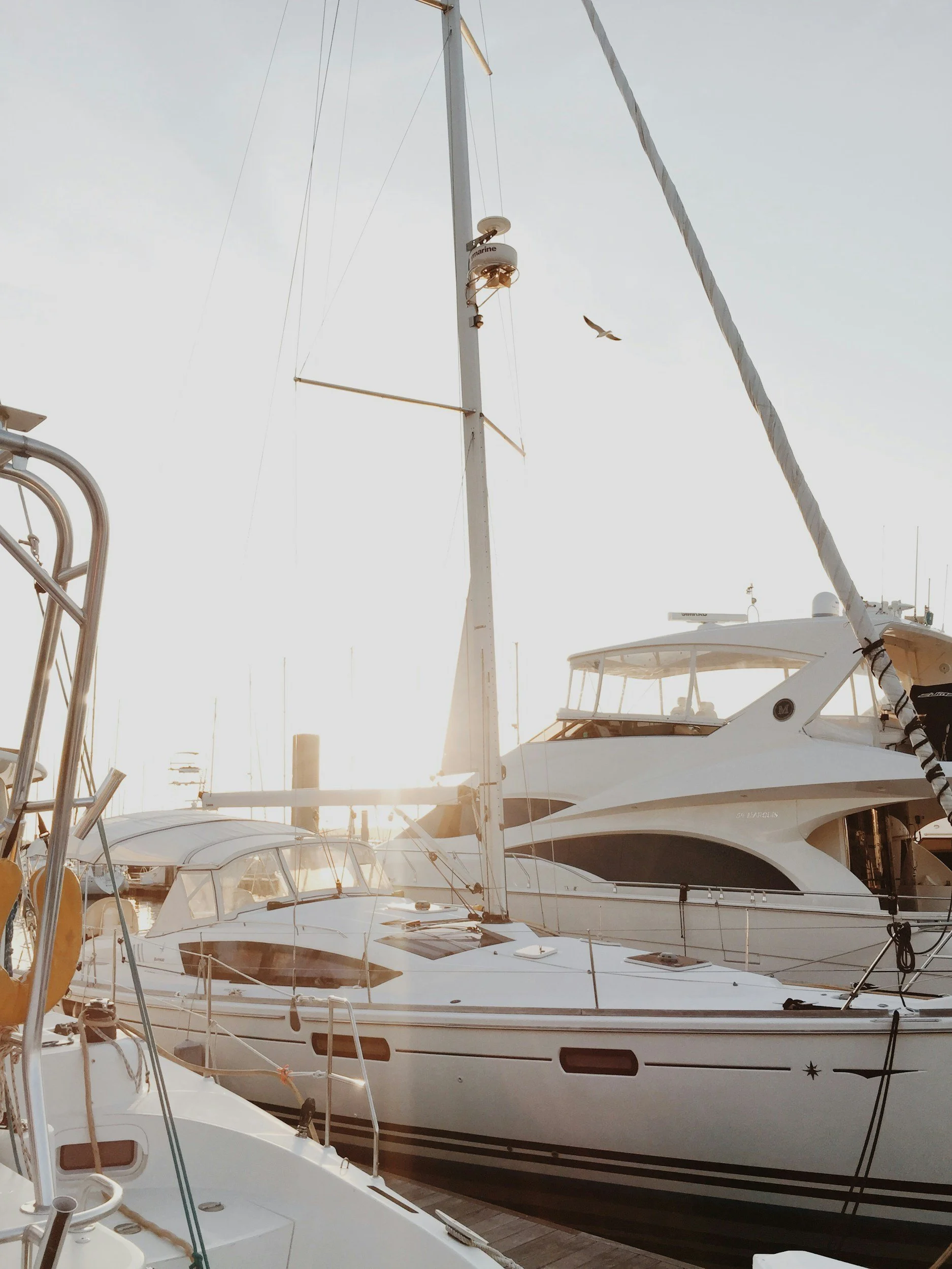 A marina with several white boats docked, including a large yacht, with the sun setting in the background.