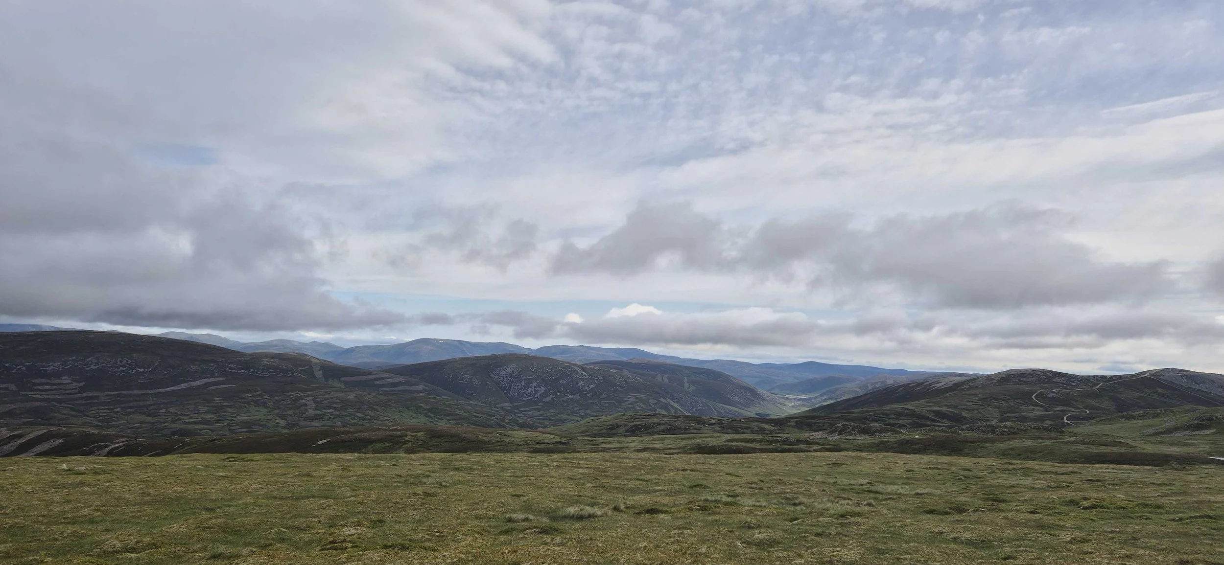 Landscape view of rolling hills and mountains under a cloudy sky with patches of blue, grassy foreground, and winding paths or roads.