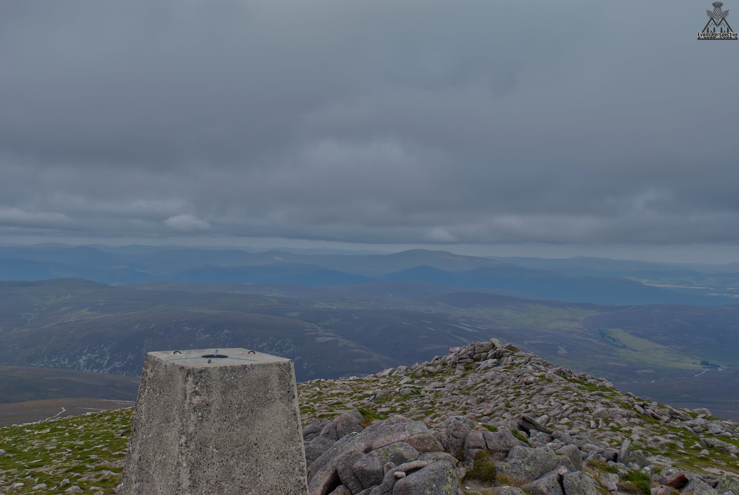 A landscape view from a mountain summit with a stone cairn in the foreground, rolling hills and distant mountains under a cloudy sky.