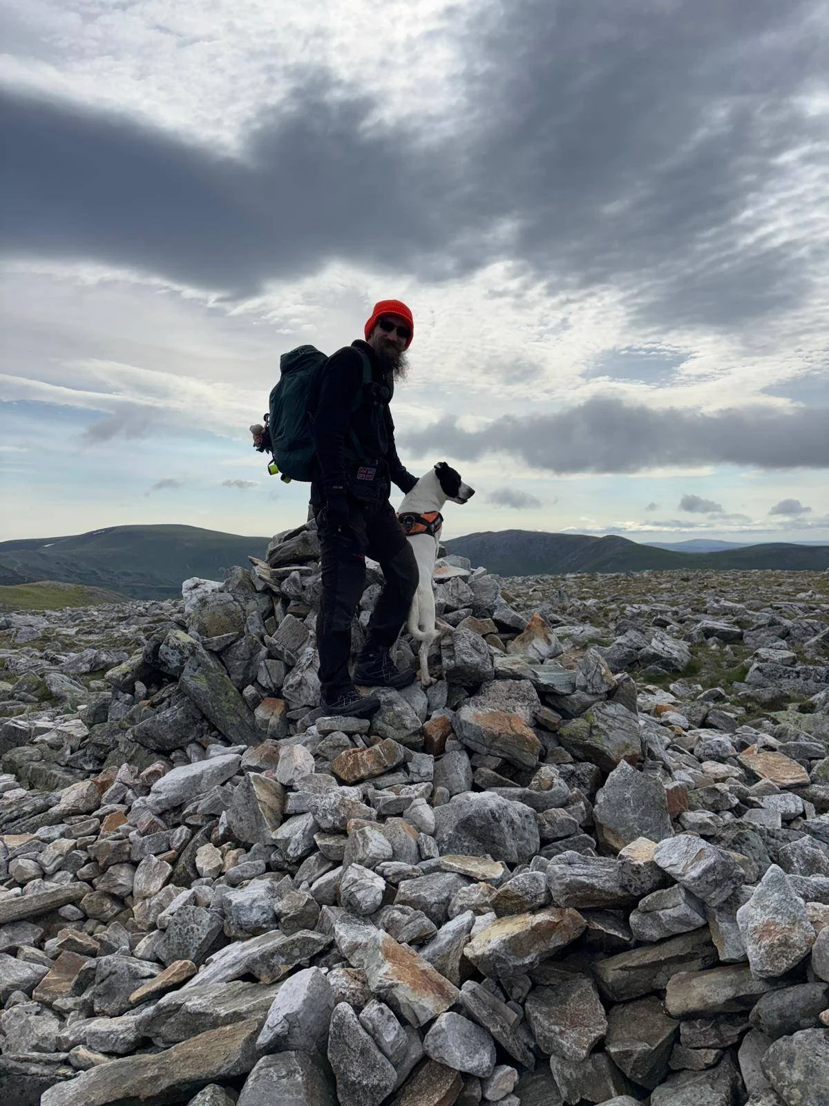 Outdoor photographer Scott standing with his dog on a rocky mountain summit cairn under dramatic skies in Scotland.