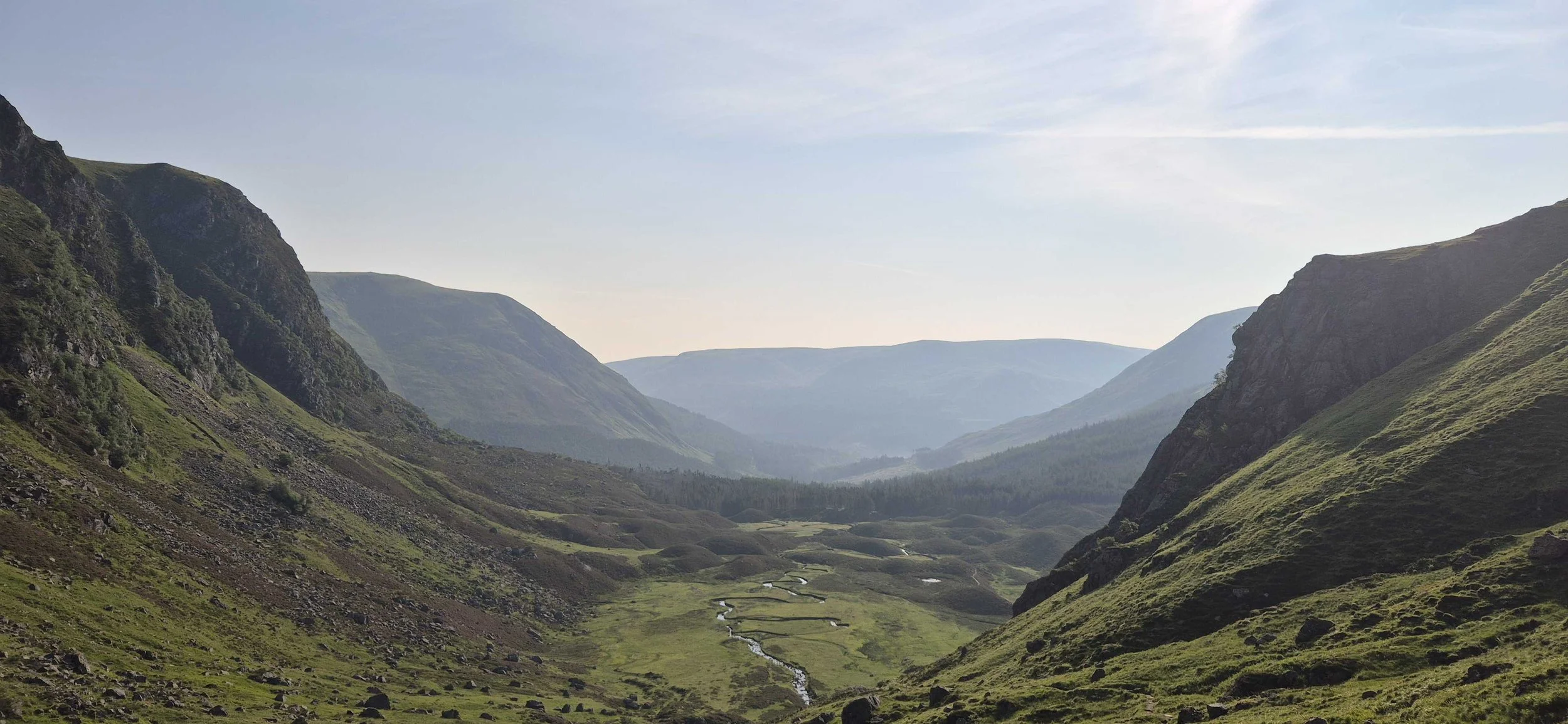 A scenic view of a deep valley with lush green hills and steep mountains on both sides, a winding river flowing through the valley, and distant mountains under a blue sky with light clouds.