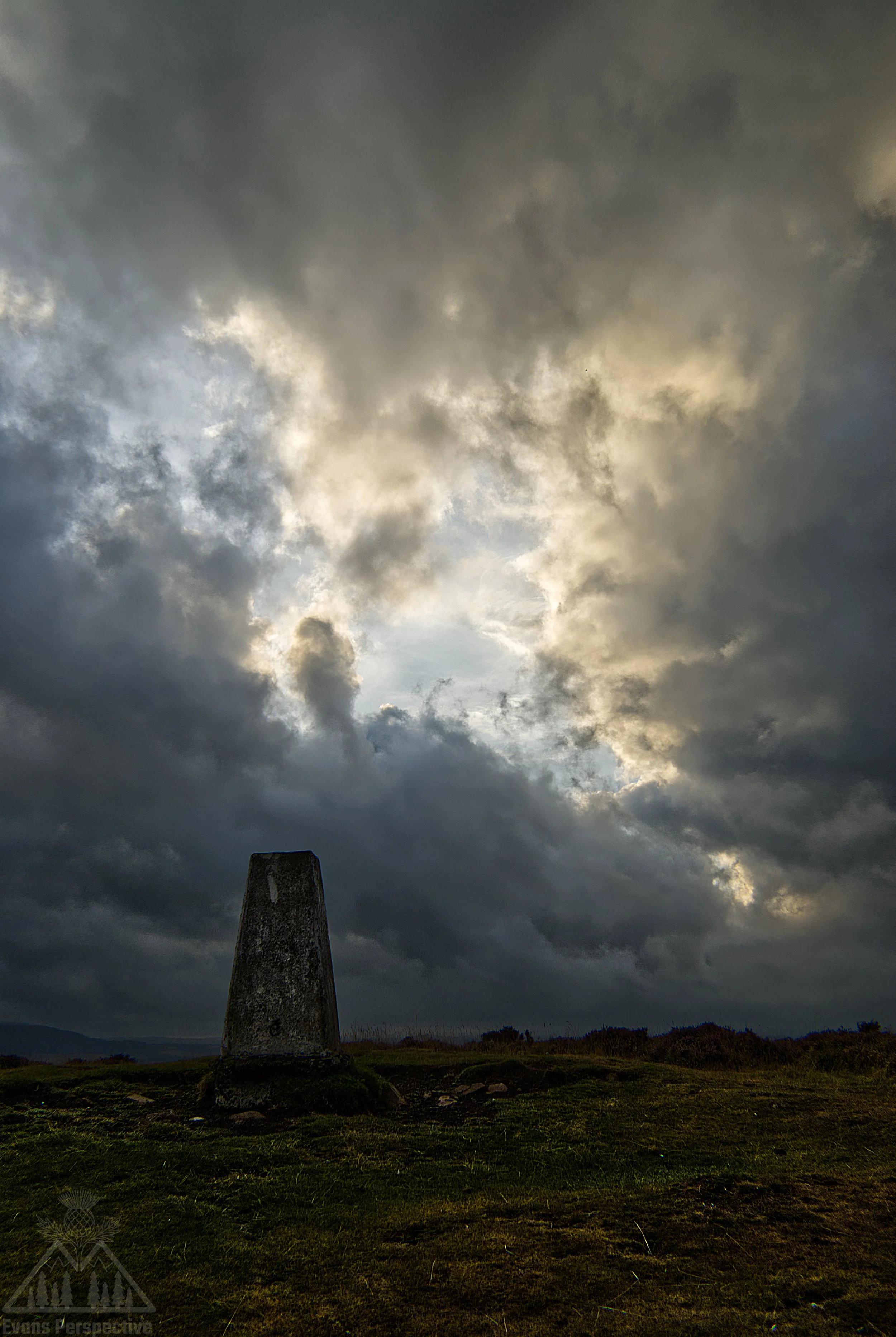 A tall, triangular stone monument standing on a grassy field under an overcast sky with dark, looming clouds.