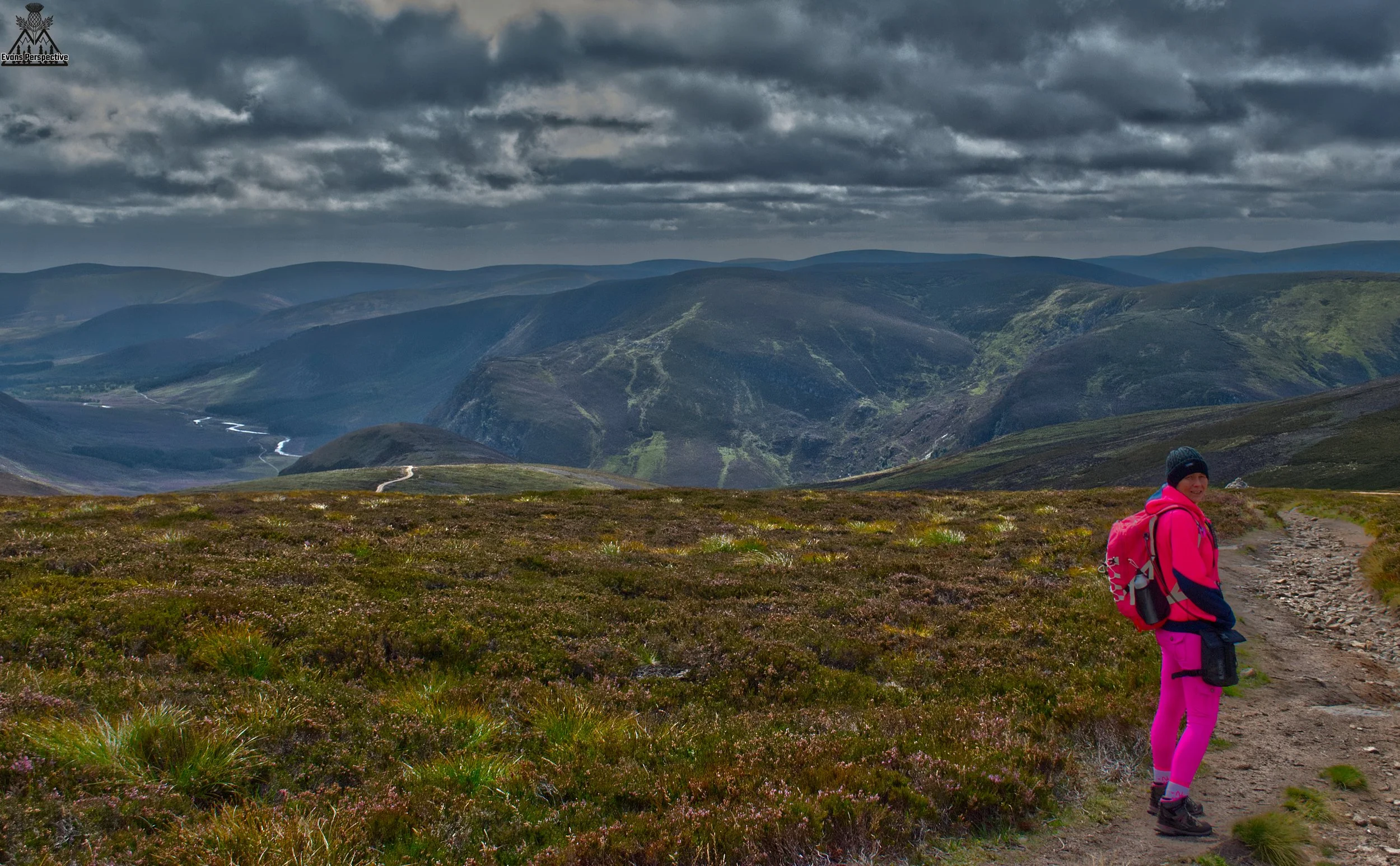 A female hiker wearing bright pink gear standing on a dirt trail looking back over a dramatic Scottish mountain landscape under heavy clouds.