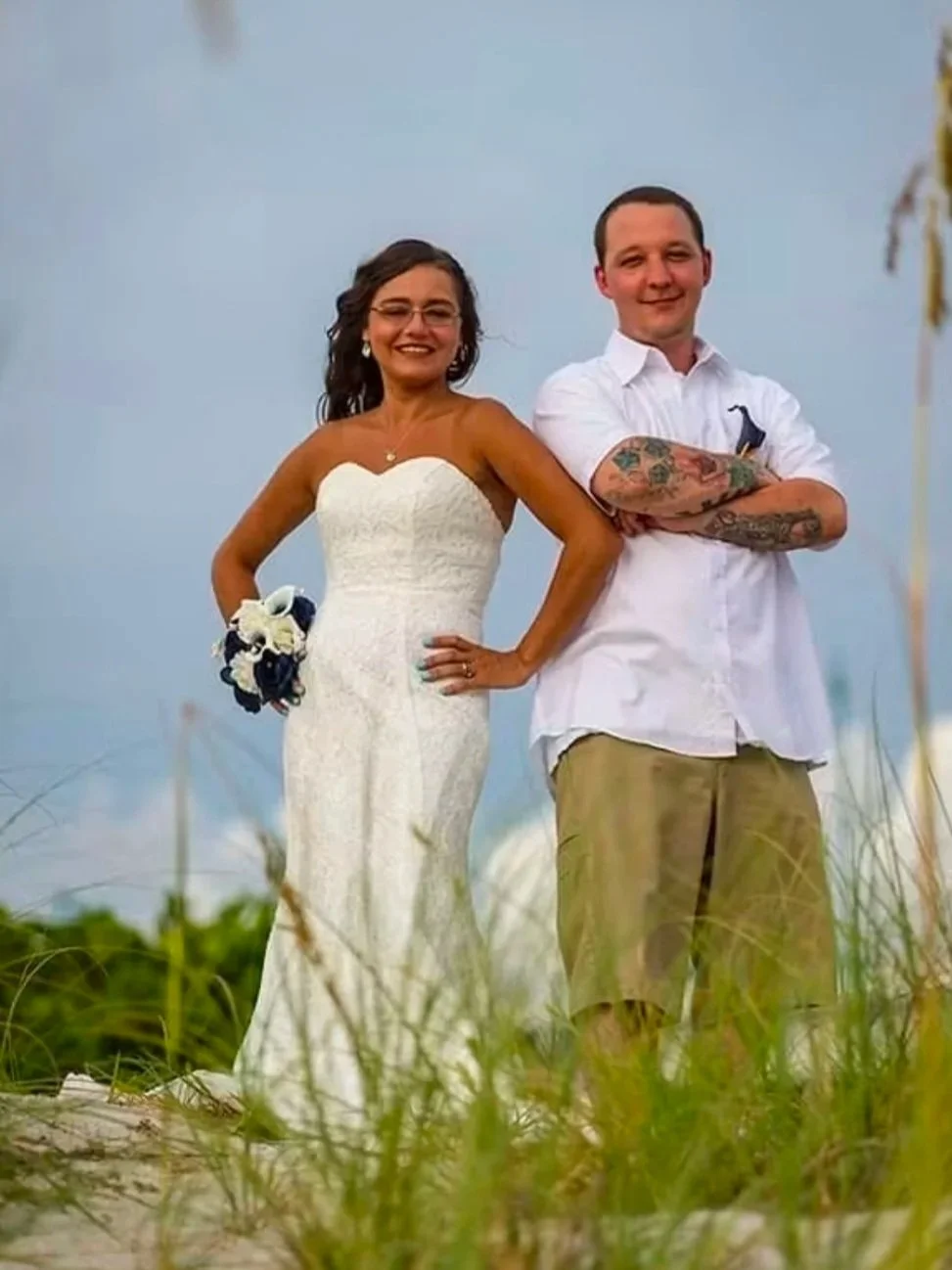 A smiling woman in a white wedding dress holding a bouquet, standing next to a man in a white shirt with tattoos on his arms, both outdoors against a blue sky background.
