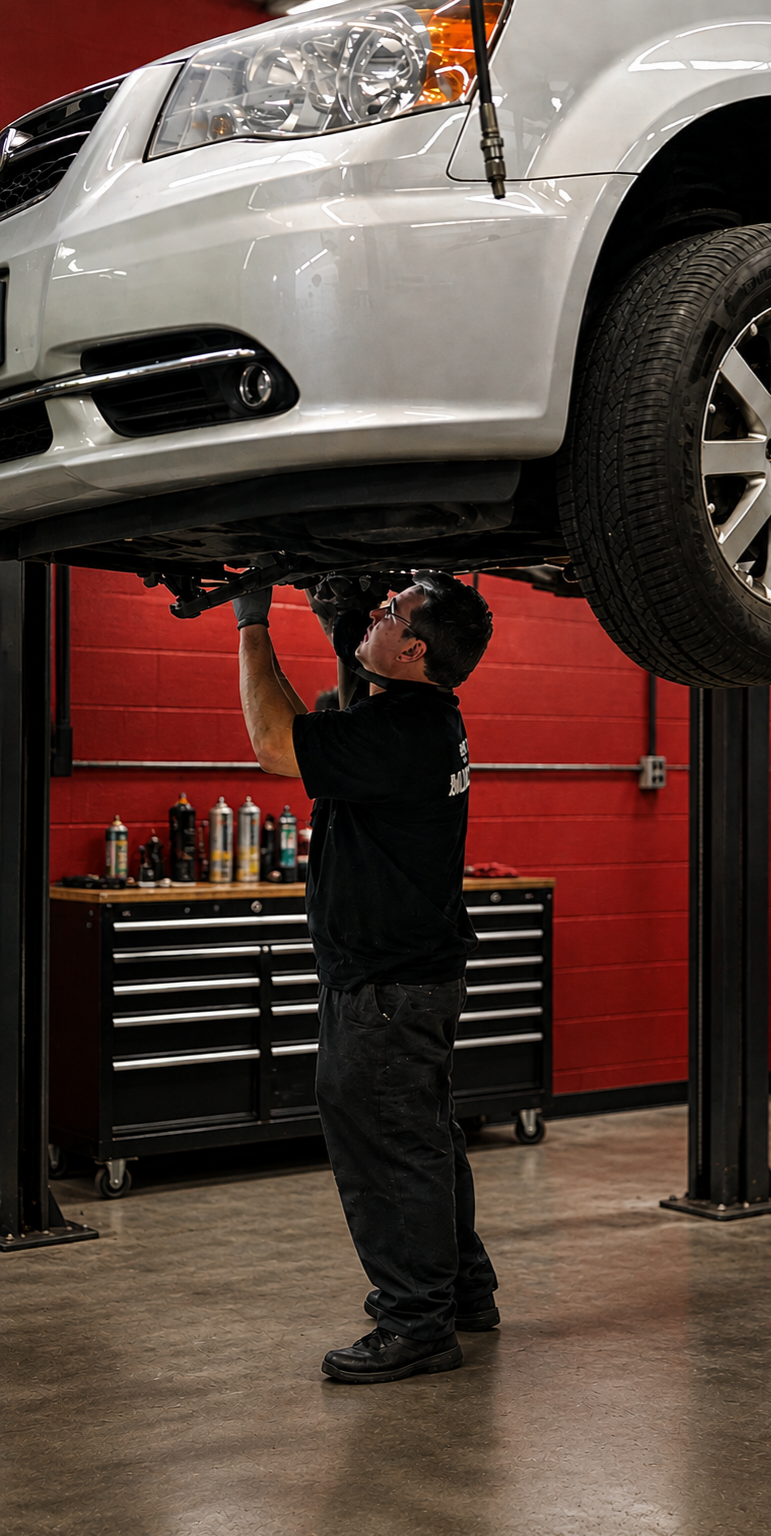 A mechanic under a white car on a lift, working on the underside of the vehicle in a garage with red walls and a workbench.