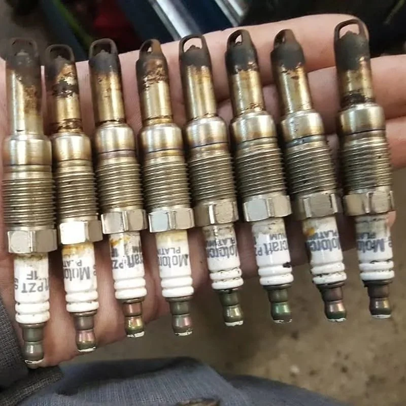 Nine spark plugs held in a person's hand, showing signs of use and carbon buildup.