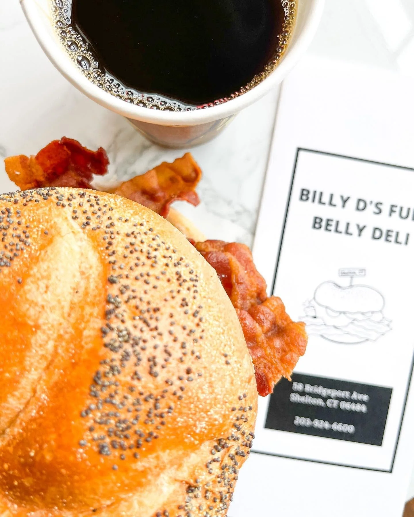 Close-up of a poppy seed bagel with a piece of crispy bacon and some orange spread, a cup of black coffee, and a menu receipt from Billy D's filled with text and a small illustration.